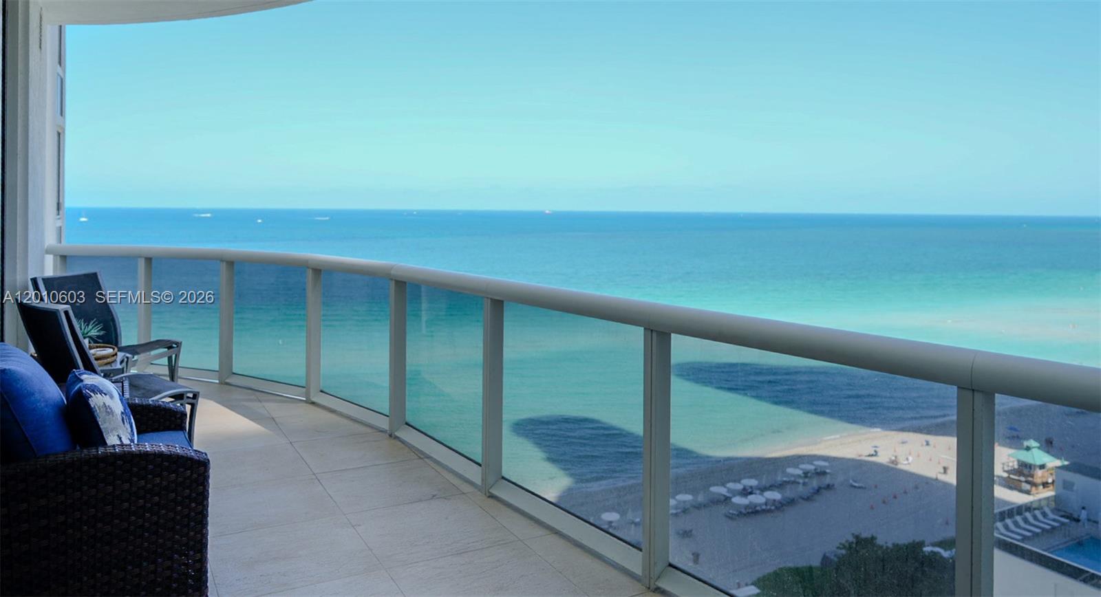 Ocean-view balcony with glass railing and seating overlooking turquoise water and a sandy beach.
