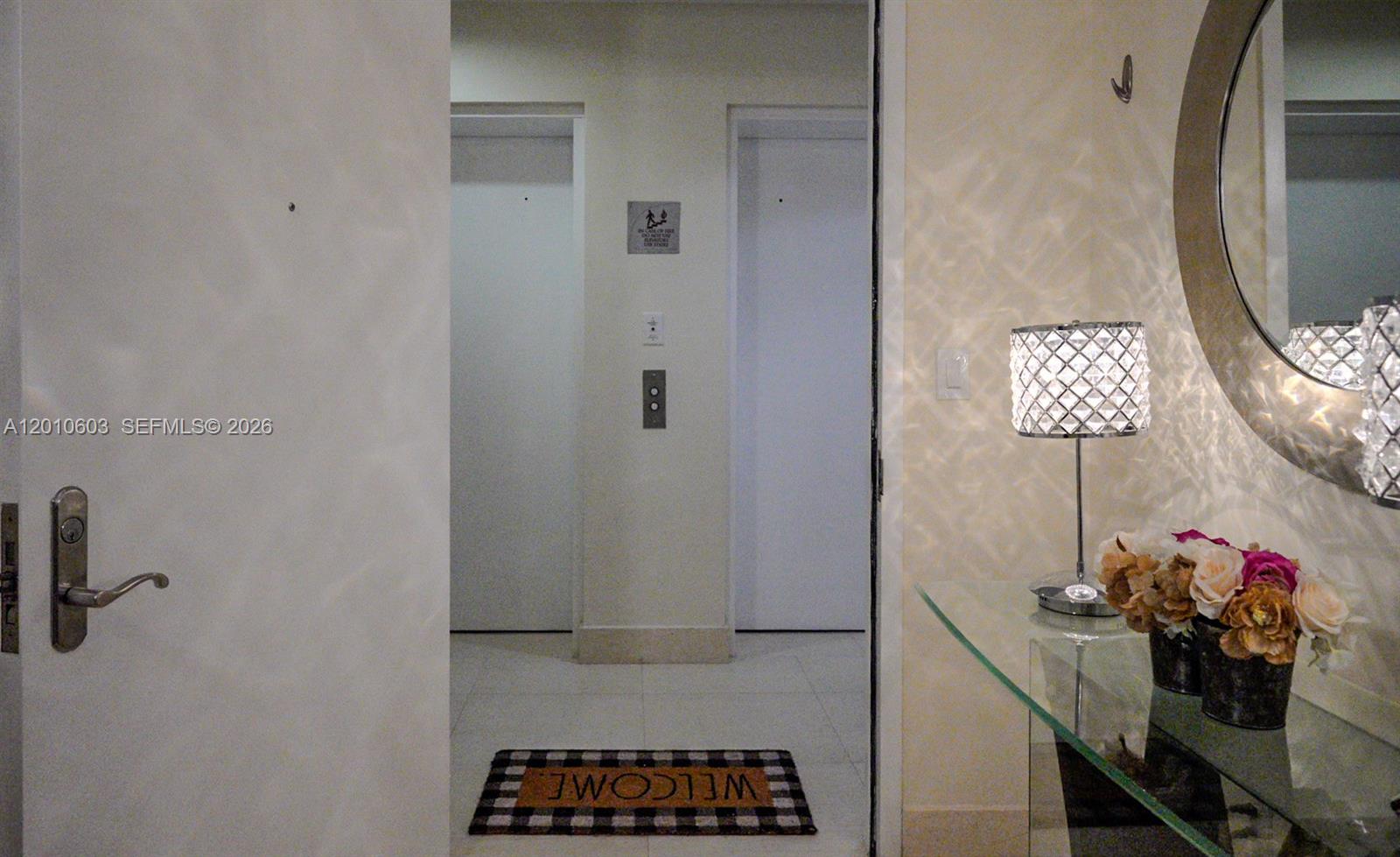 Condo entry foyer with elevator doors, a glass console table, flowers, and a round mirror.