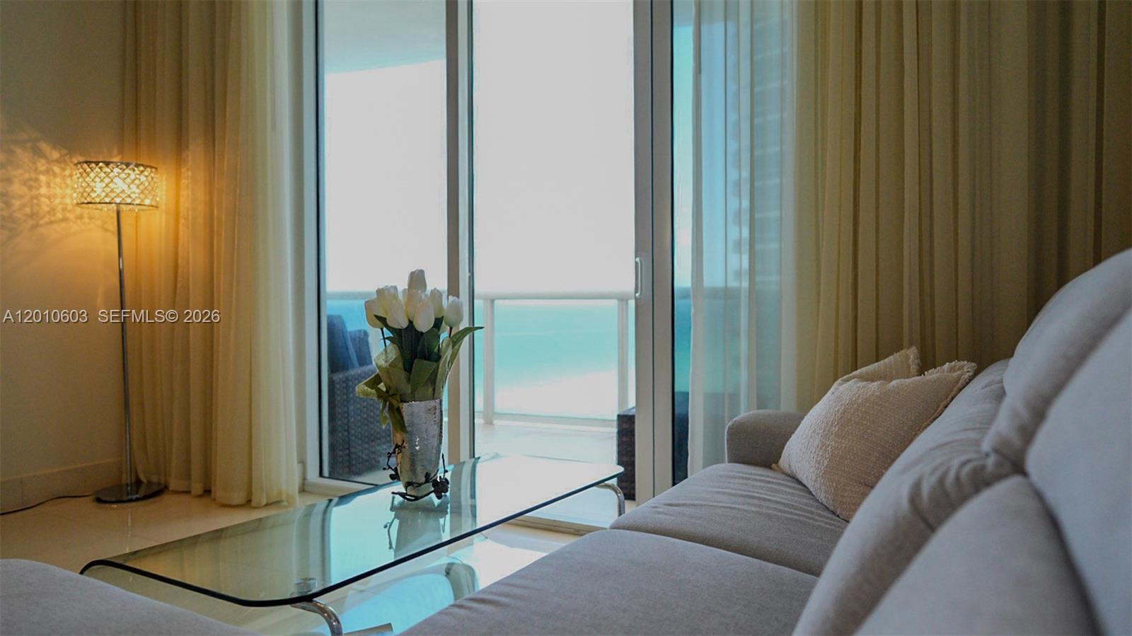 Living room with a gray sectional and glass coffee table overlooking the ocean through sliding doors.