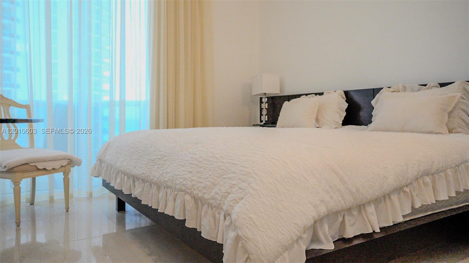 Bedroom with a large white quilted bed beside bright curtained windows.