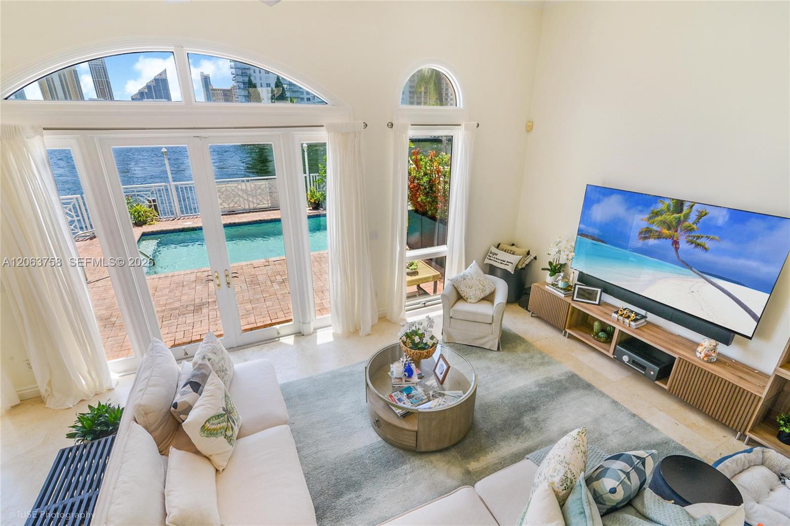 Furnished living room with arched windows overlooking a pool and waterfront.