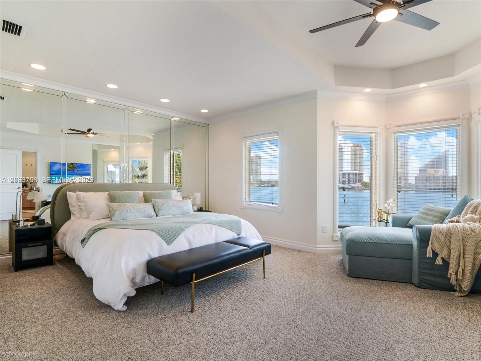 Waterfront bedroom with mirrored panels, a pale blue chaise lounge, and windows overlooking the bay and skyline.