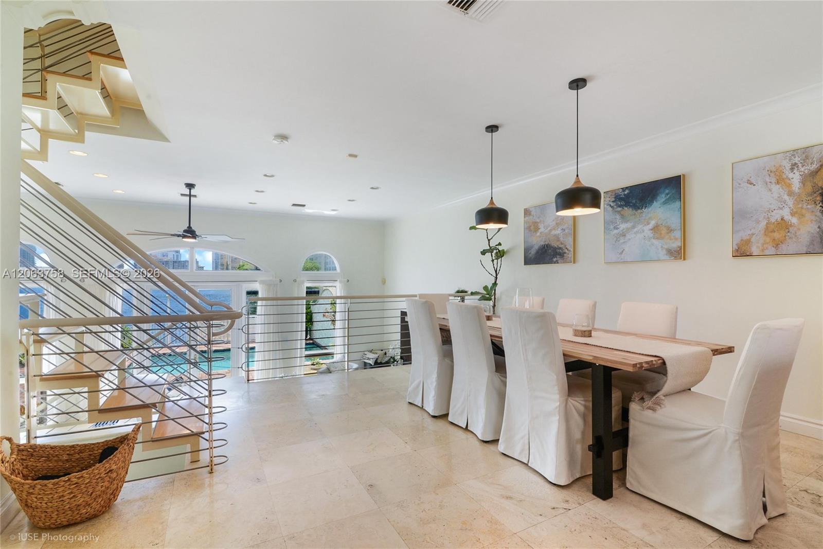 Dining area with a long wood table, white chairs, and arched windows overlooking water.