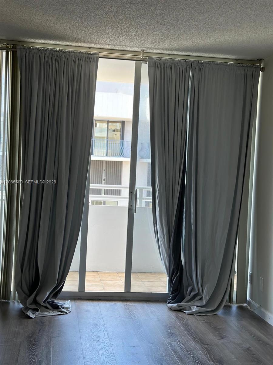 Vacant condo room with gray curtains, wood-look flooring, and sliding glass doors to a tiled balcony.