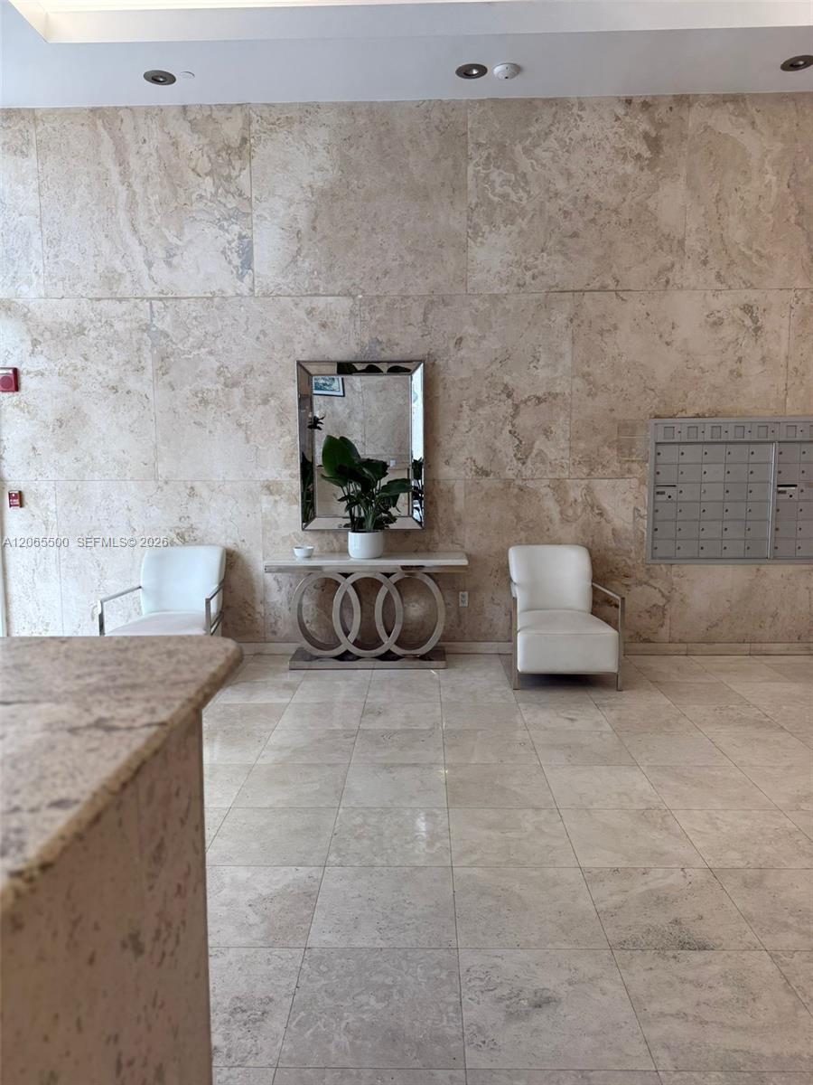Condominium lobby with beige stone walls, white chairs, a mirrored console, and wall-mounted mailboxes.