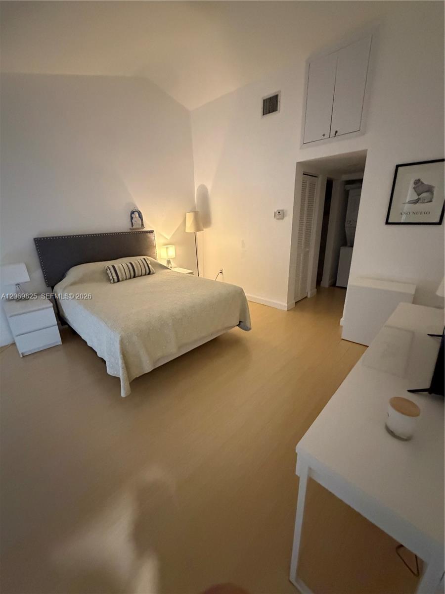 Furnished bedroom with a queen bed, dark upholstered headboard, and light wood flooring.