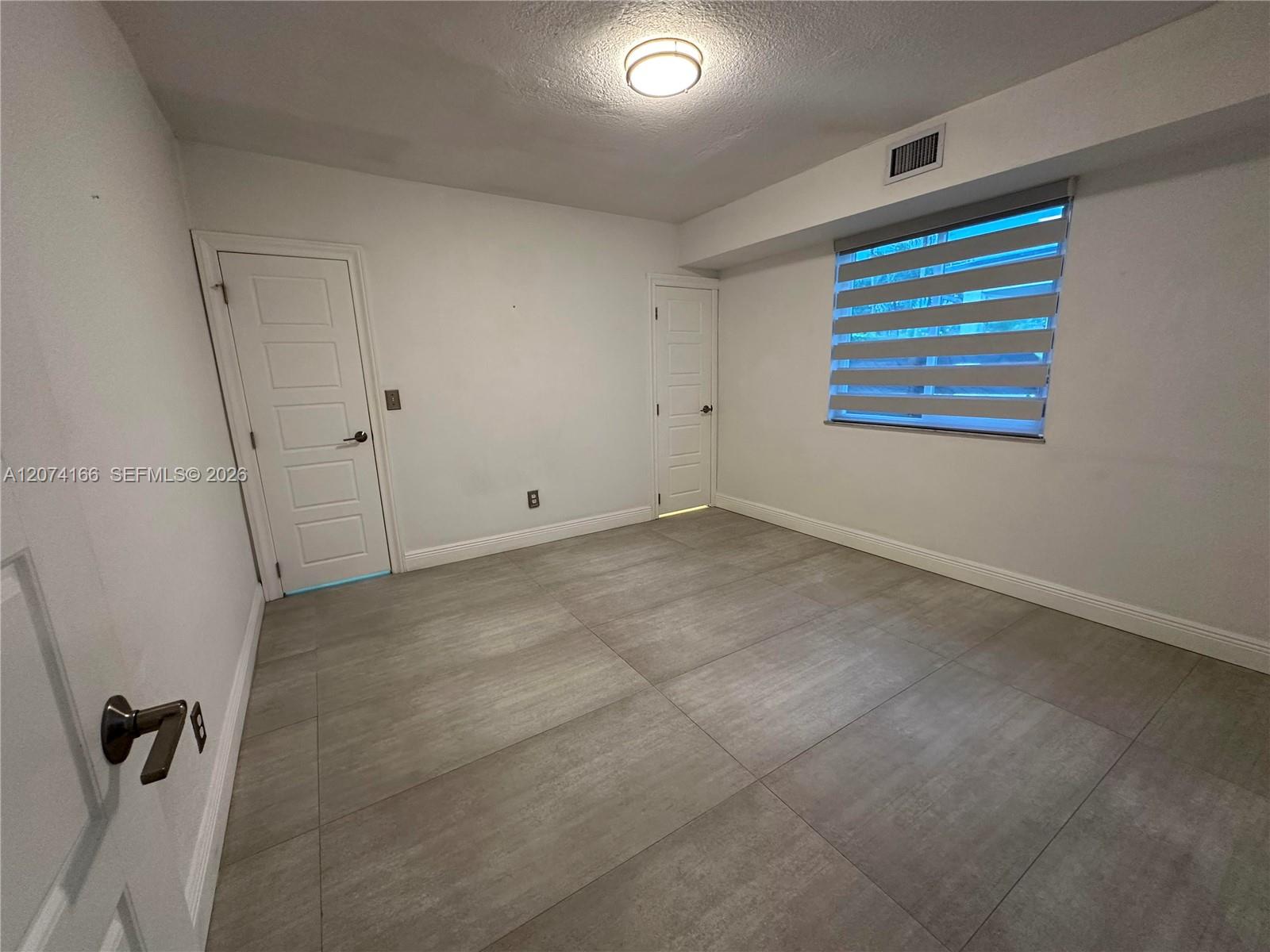 Empty tiled room with two white doors and a window with zebra blinds.