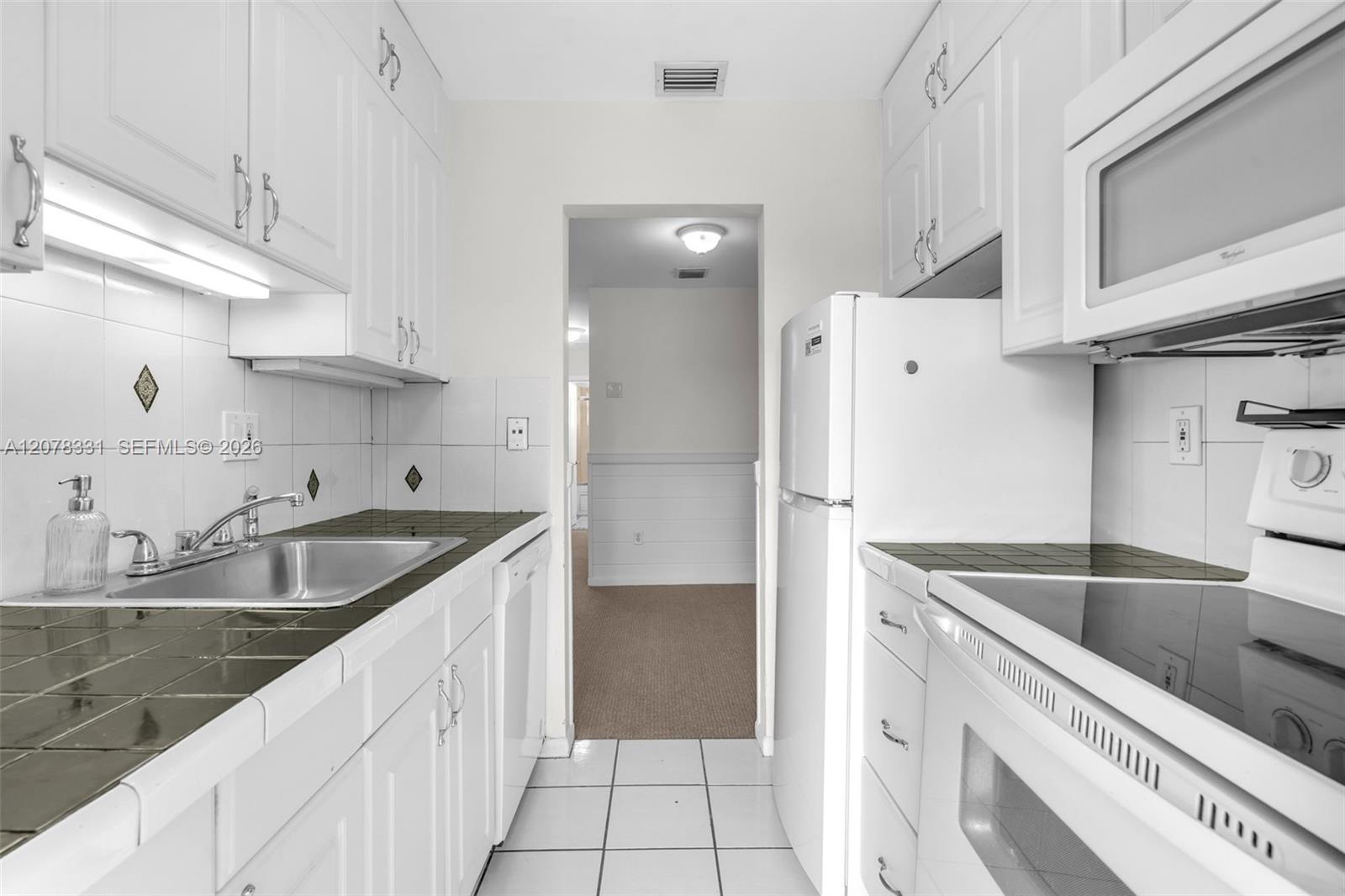 Galley kitchen with white cabinets, dark green tiled counters, stainless steel sink, and white appliances.