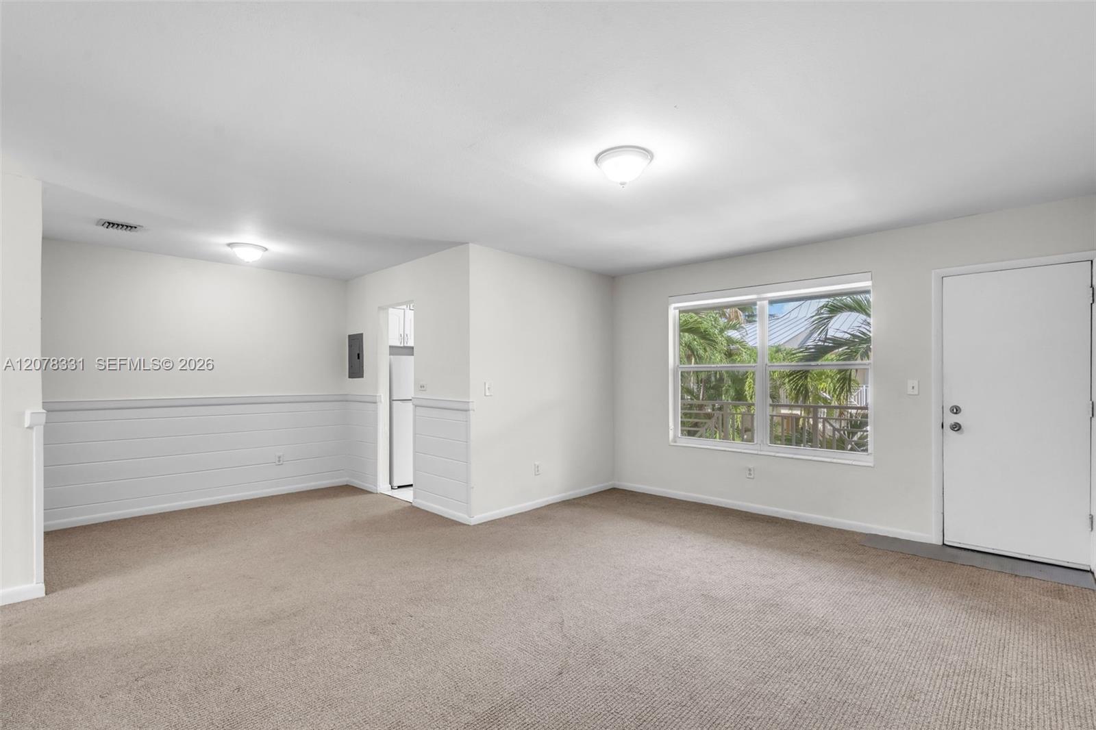 Vacant living room with beige carpet, white paneled walls, and a window overlooking palm trees.