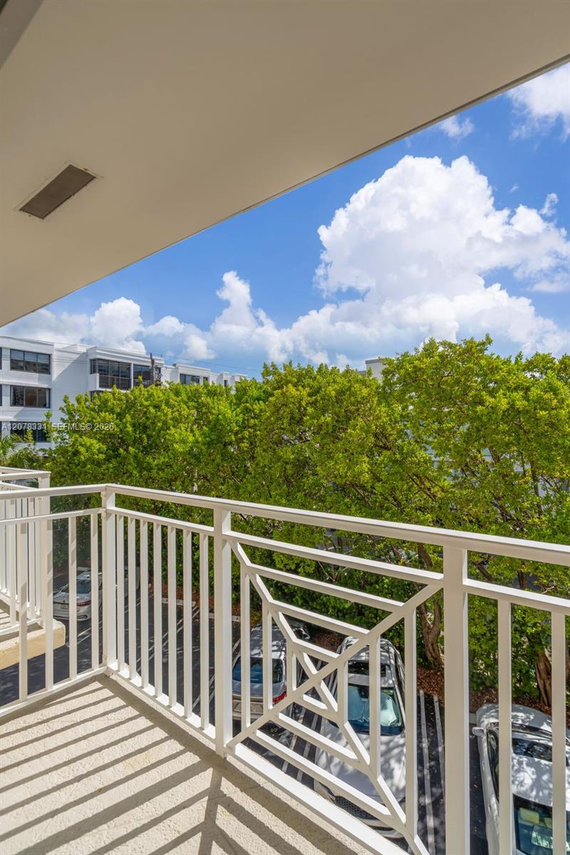 Covered apartment balcony with white cross-pattern railings overlooking trees and parked cars.