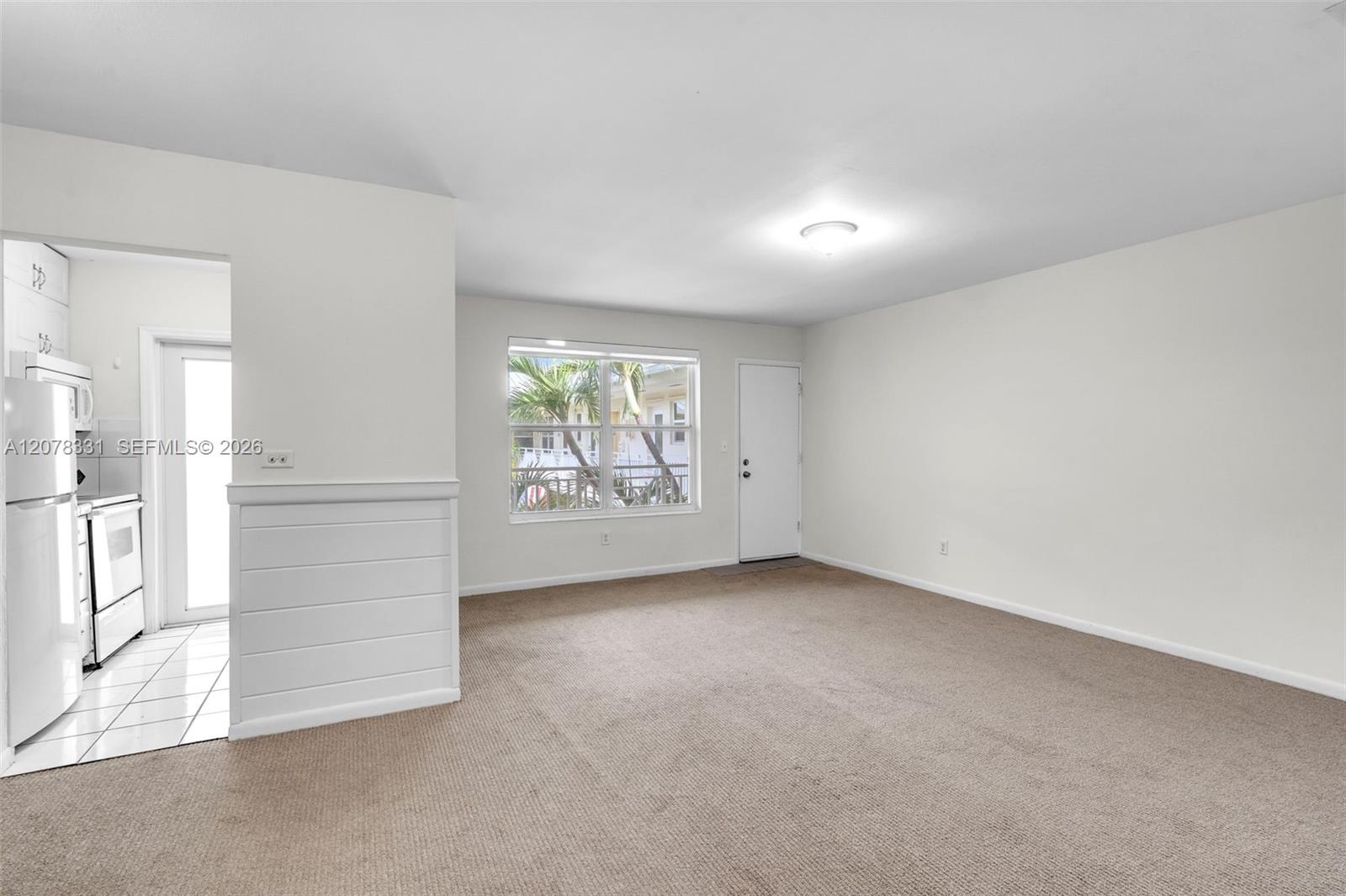 Vacant living room with beige carpet, a window overlooking palms, and an adjoining kitchen.
