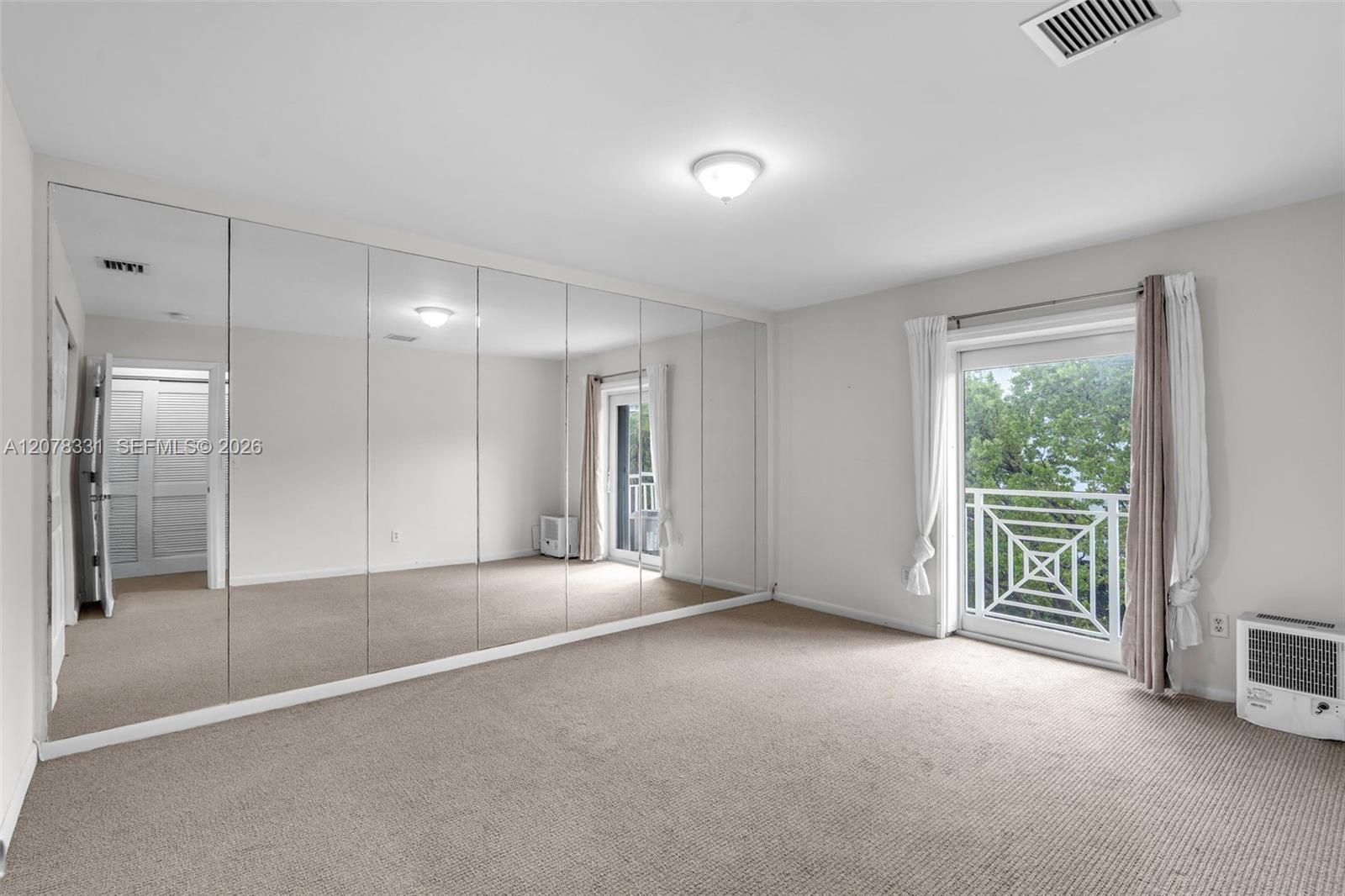 Empty carpeted bedroom with mirrored closet doors and a balcony door overlooking trees.