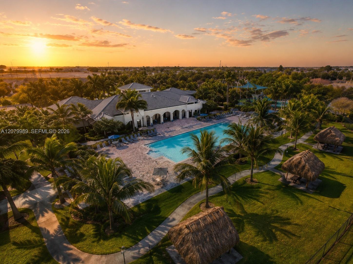 Aerial view of a community pool and clubhouse surrounded by palm trees and thatched cabanas at sunset.
