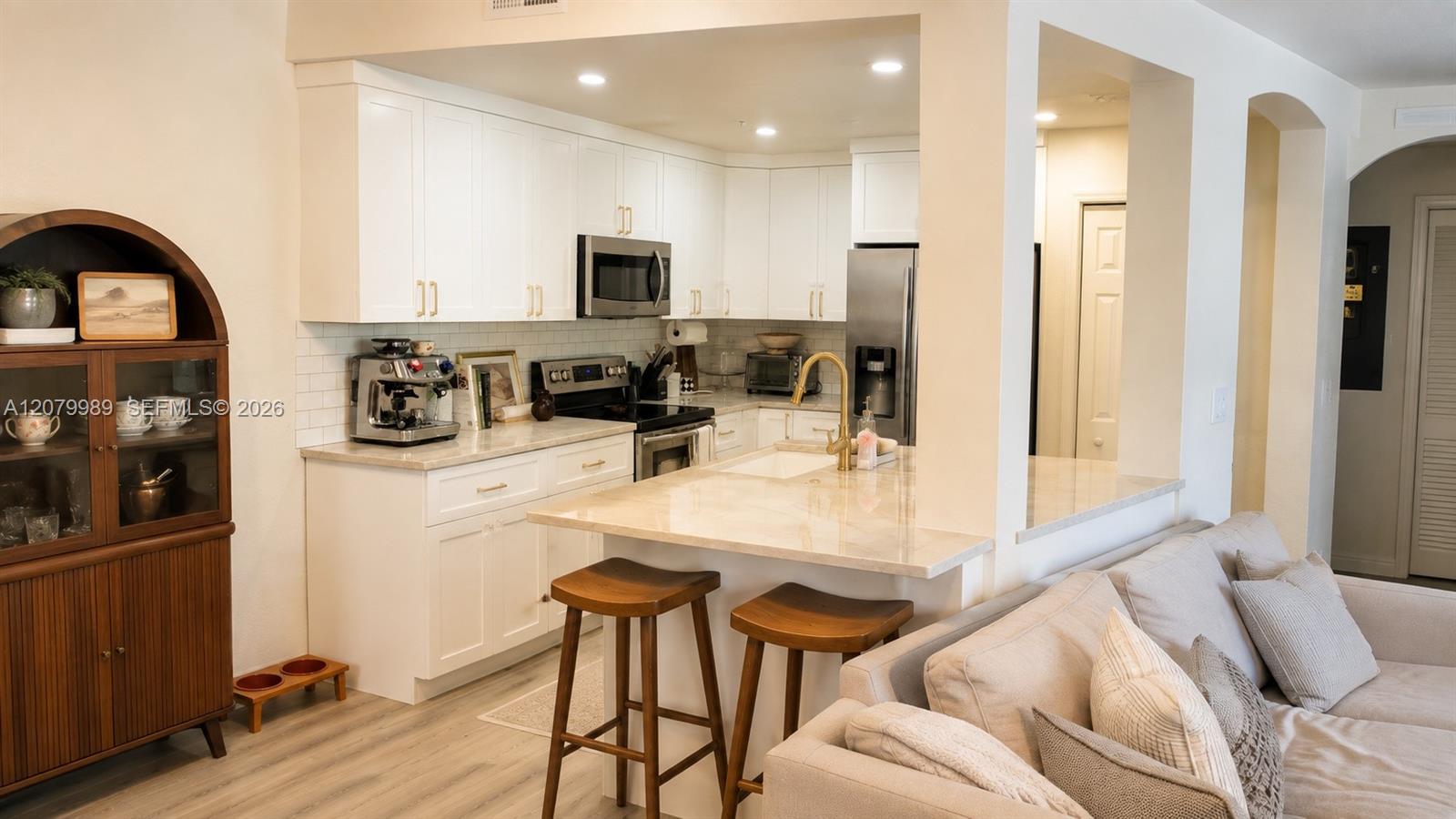 Open-plan kitchen with white cabinets, stainless steel appliances, and a marble-look island beside a light sofa.