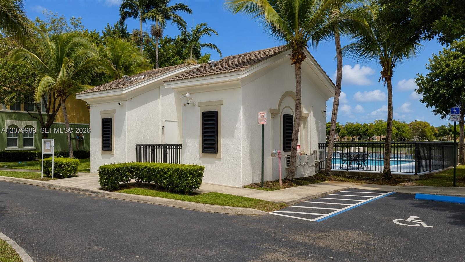 White stucco community building beside a fenced swimming pool, with palm trees and accessible parking.