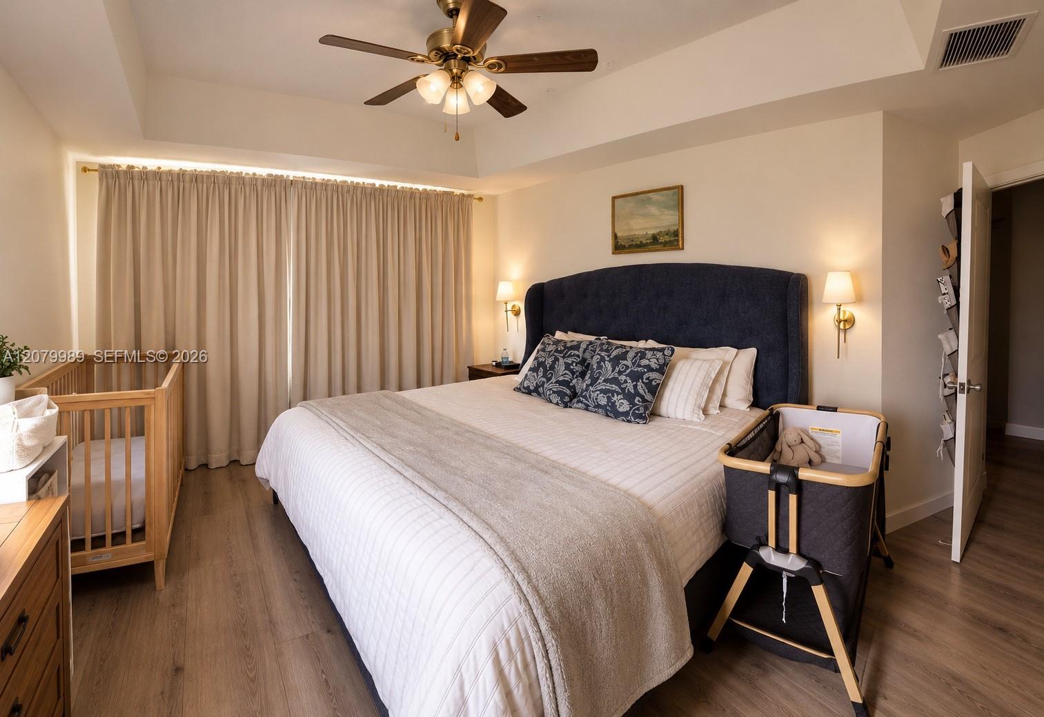 Furnished bedroom with a king bed, dark upholstered headboard, wooden crib, and baby bassinet.