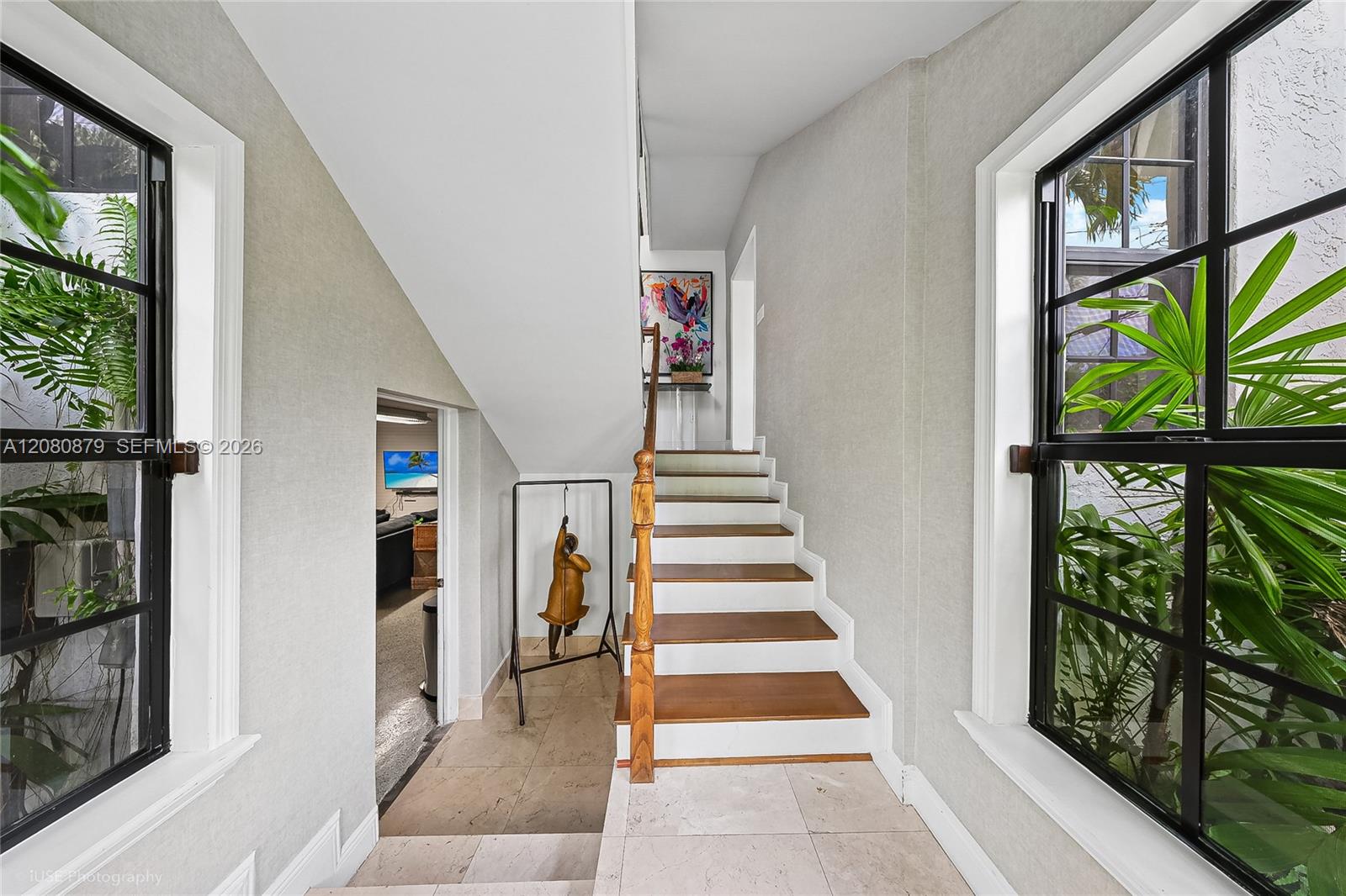 Entry hall with a wood-trimmed staircase, black-framed windows, and tropical greenery outside.