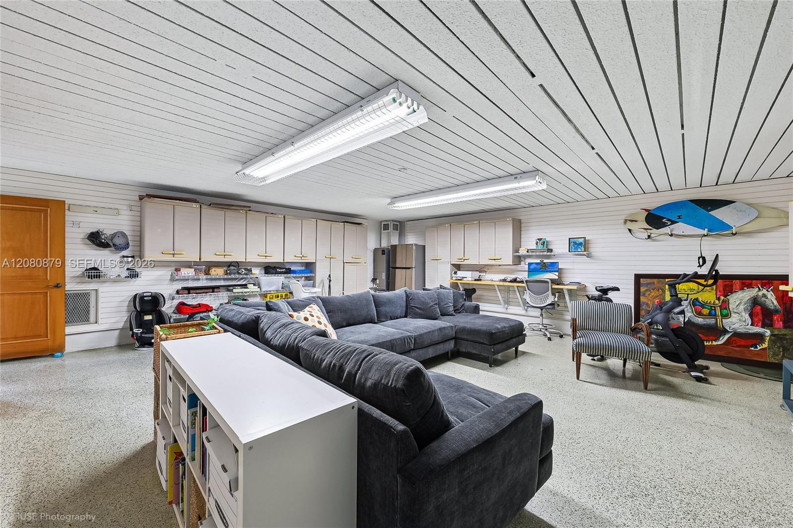 Multipurpose room with a large gray sectional, wall cabinets, desk, and surfboards mounted above colorful artwork.