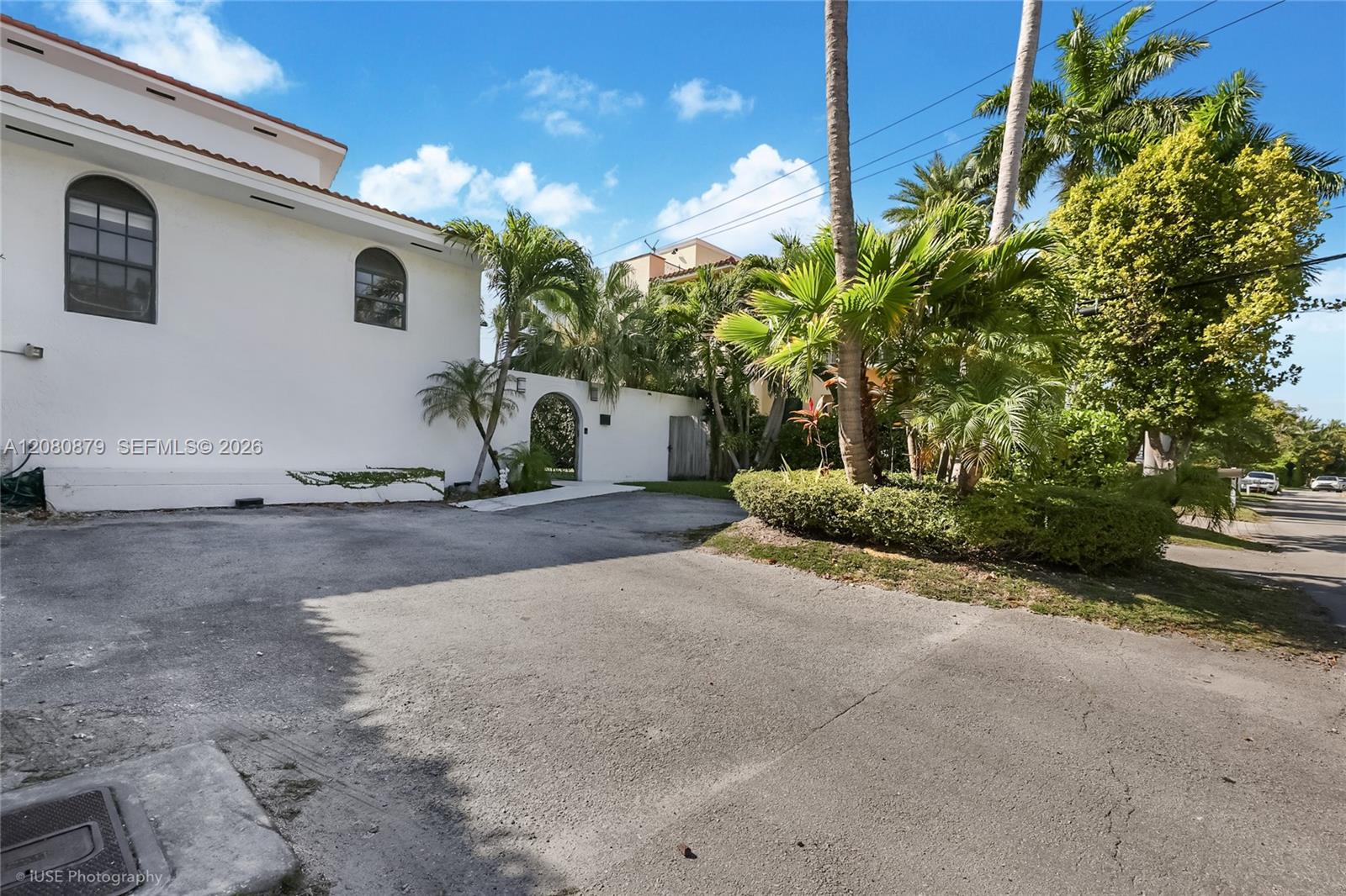 White two-story building with arched windows, a paved driveway, and lush tropical landscaping.