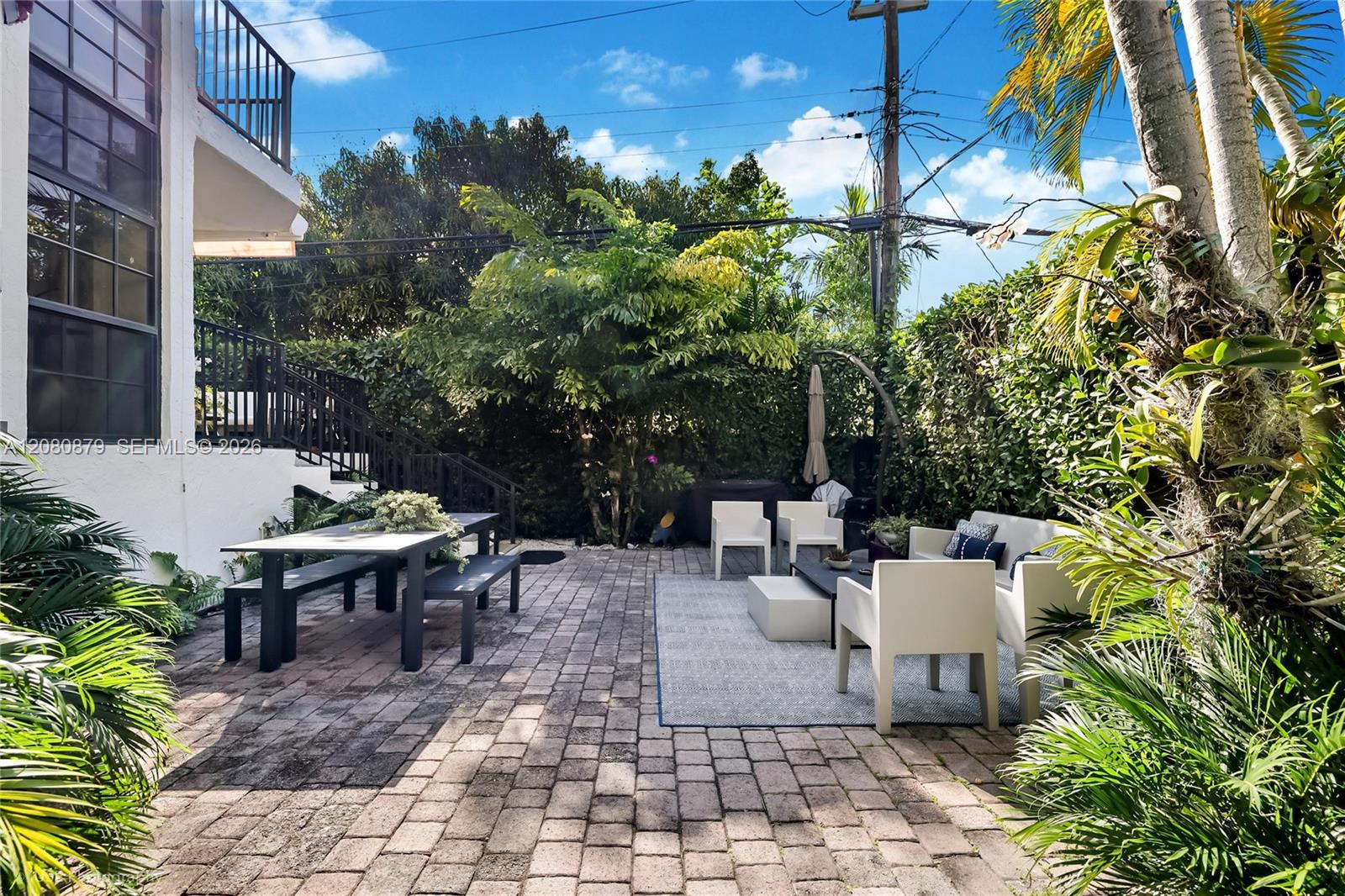 Tropical patio with paver flooring, white lounge furniture, and a dining table beside the home.