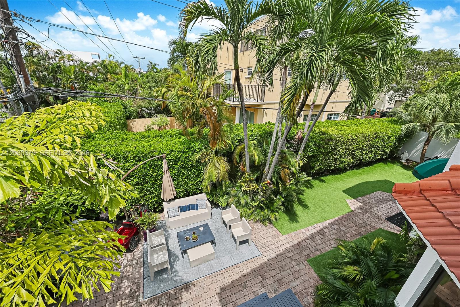 Elevated backyard patio with outdoor seating, artificial turf, pavers, and tropical landscaping.