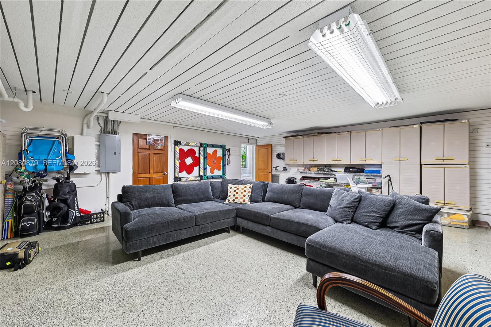 Furnished garage with a large gray sectional, wall of storage cabinets, golf clubs, and a basketball hoop.
