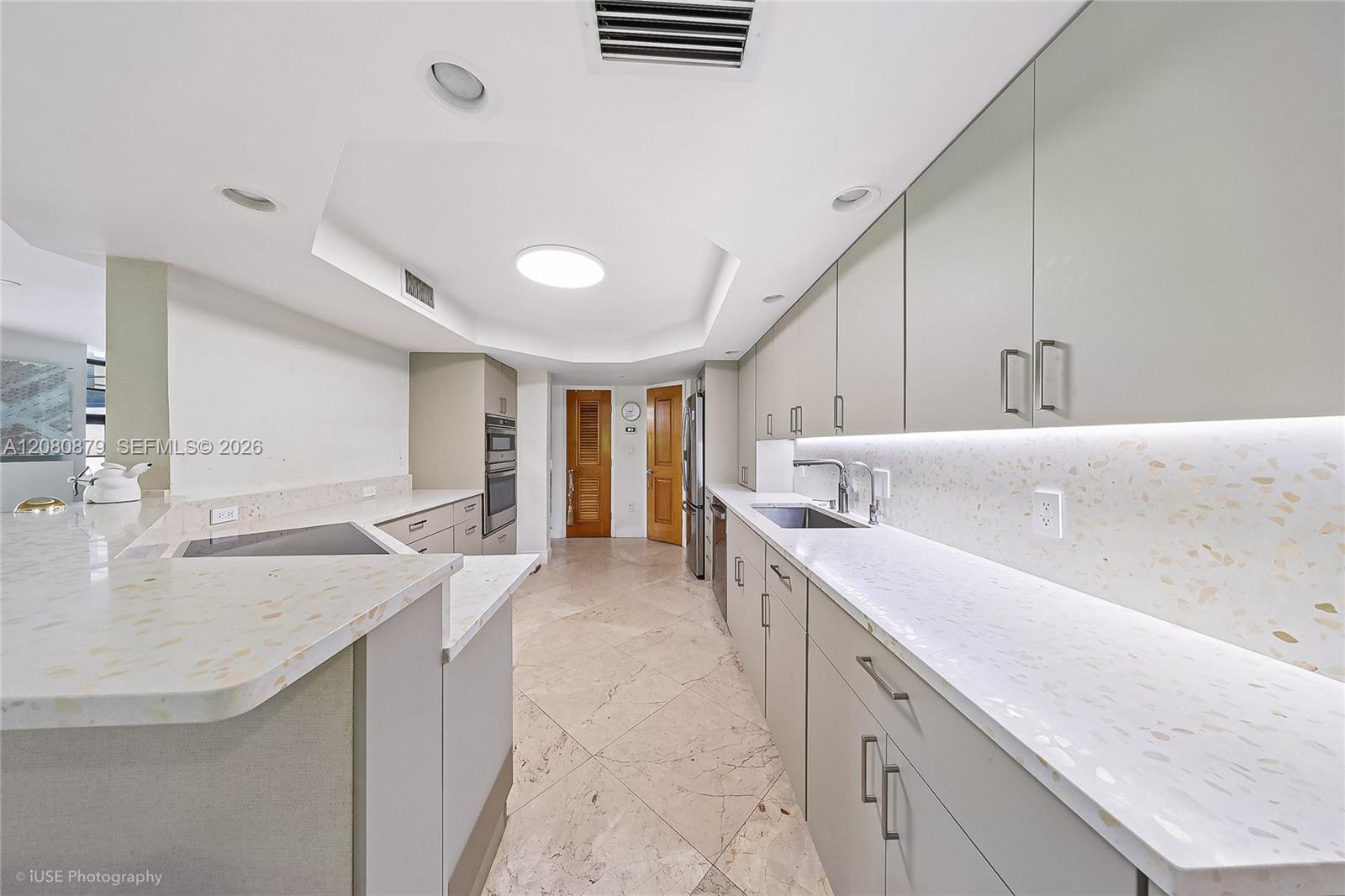 Kitchen with pale stone counters, extensive cabinetry, a peninsula, and built-in ovens.
