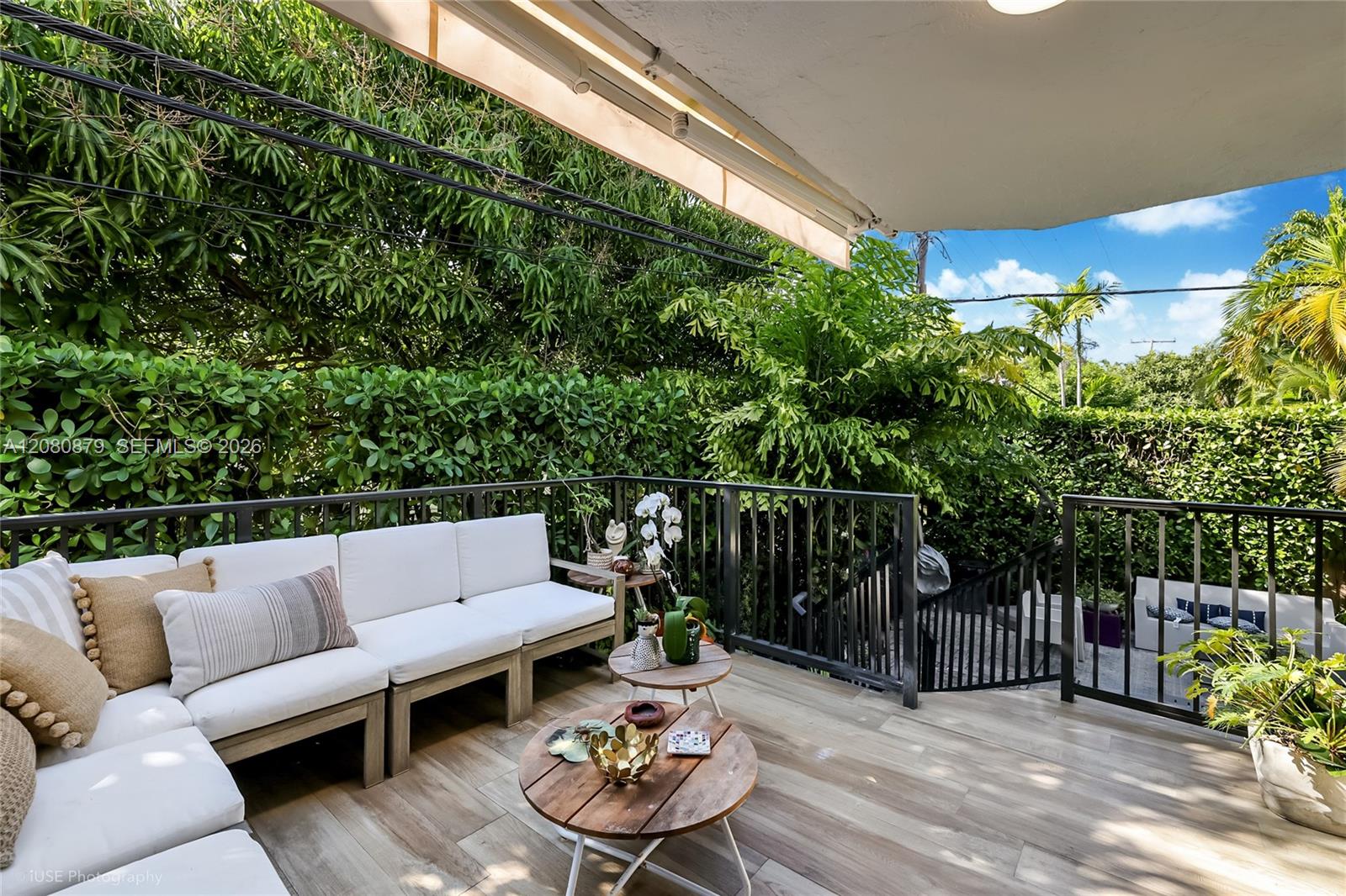 Covered patio with a white sectional, wooden coffee tables, and dense tropical greenery beyond the railing.