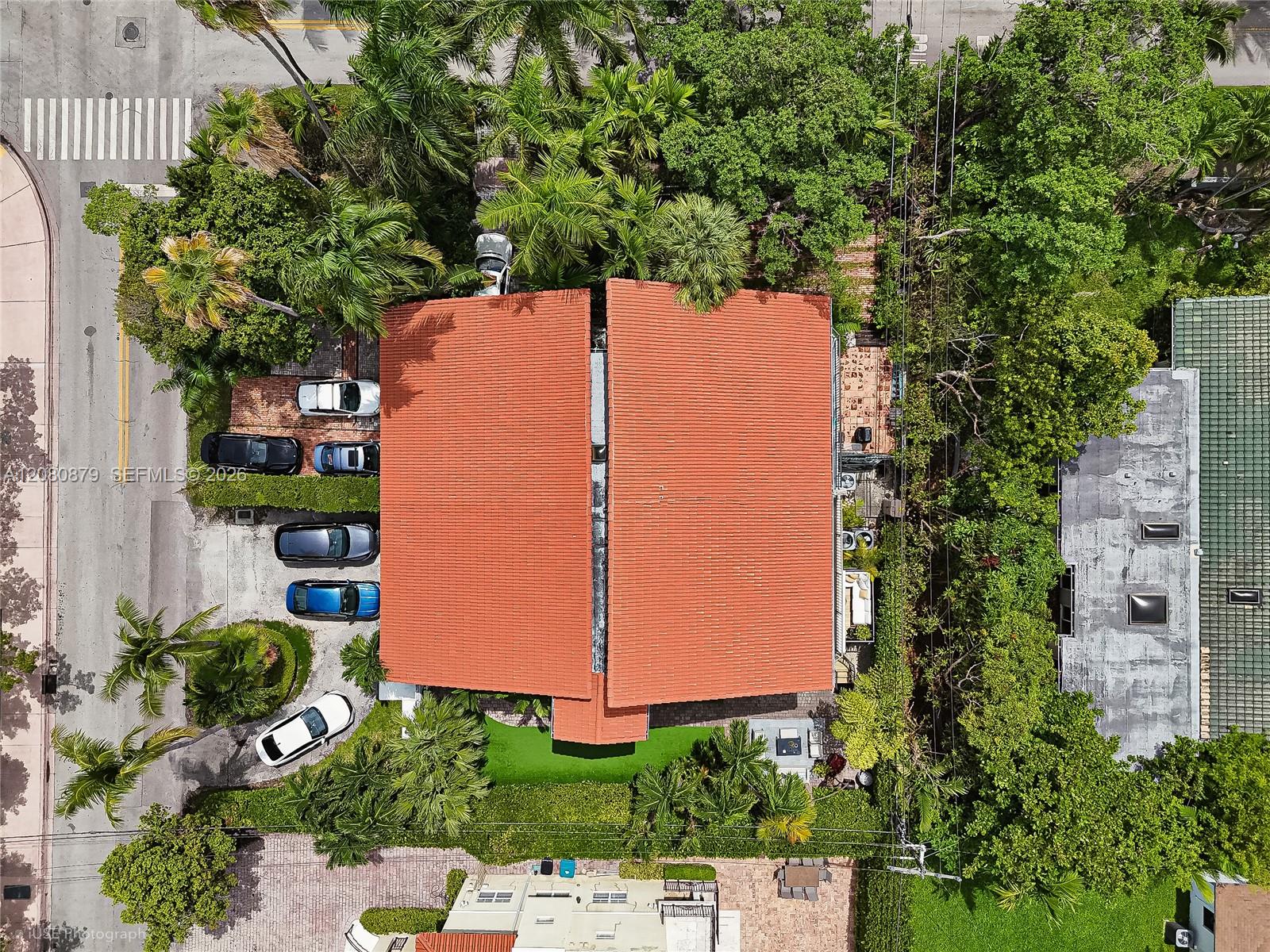 Aerial view of a red-roofed home surrounded by palms, with a driveway and parked cars.