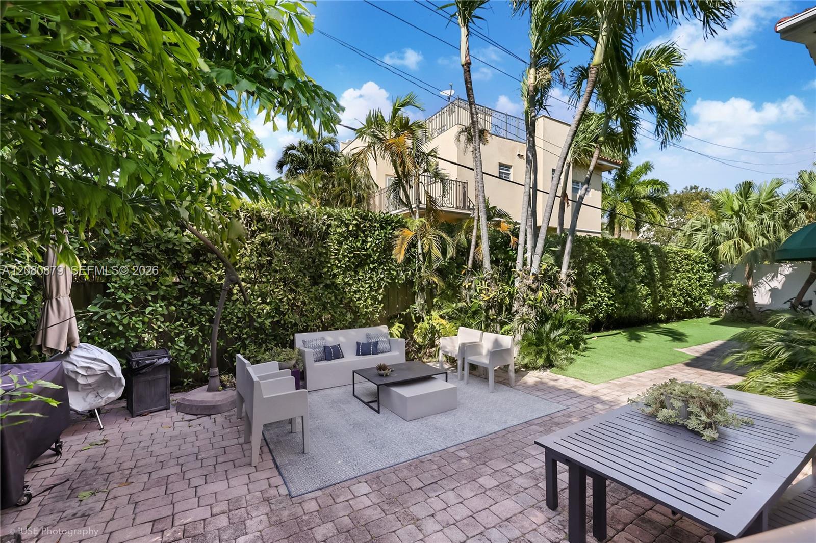 Tropical backyard patio with gray lounge seating, palms, hedges, and a paved lawn path.