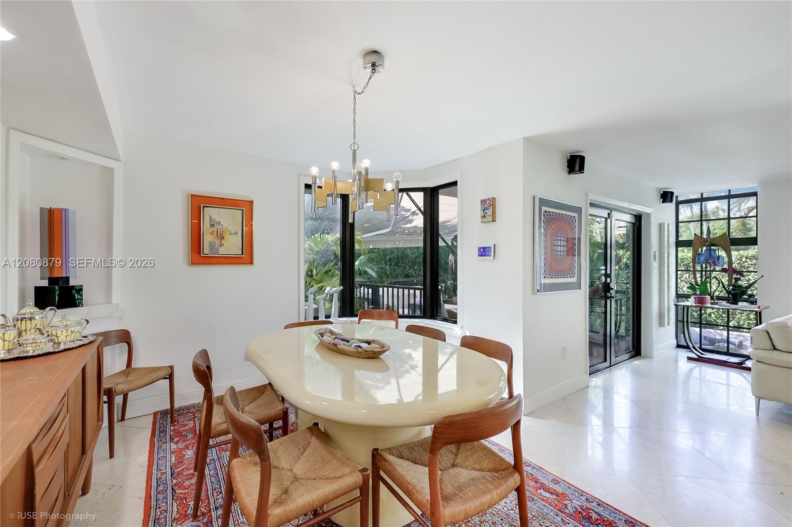 Dining room with an oval table, woven chairs, patterned rug, and glass doors overlooking greenery.
