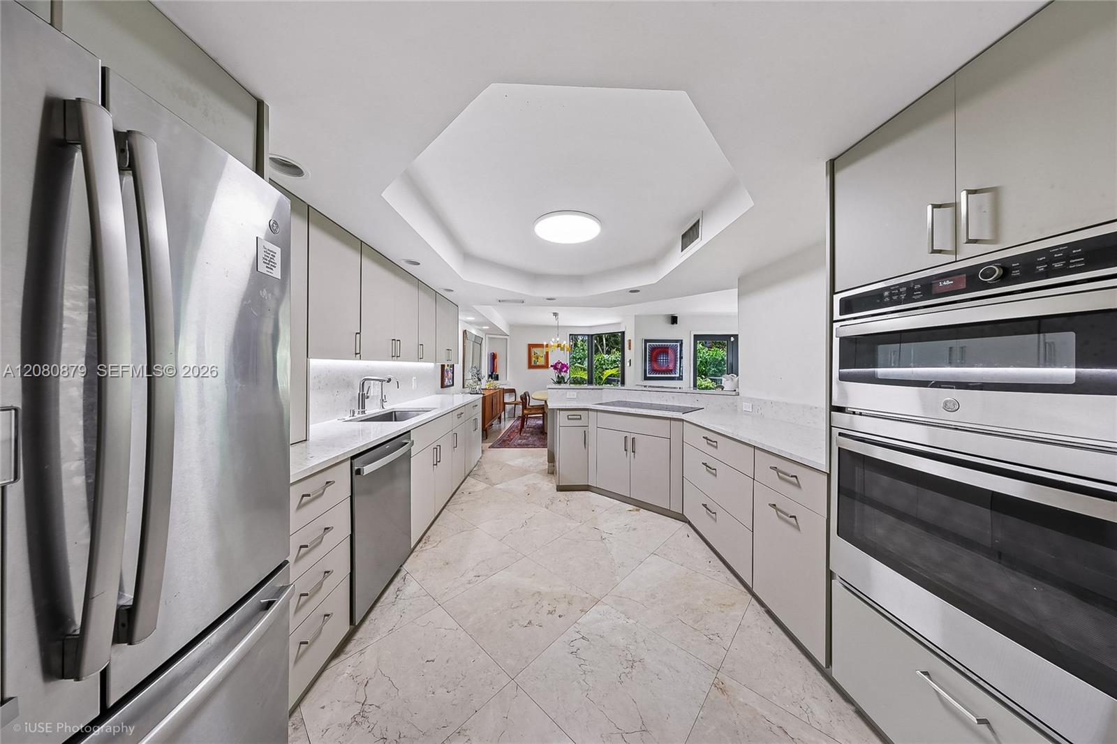 Modern white kitchen with stone counters, stainless appliances, and a cooktop island overlooking greenery.