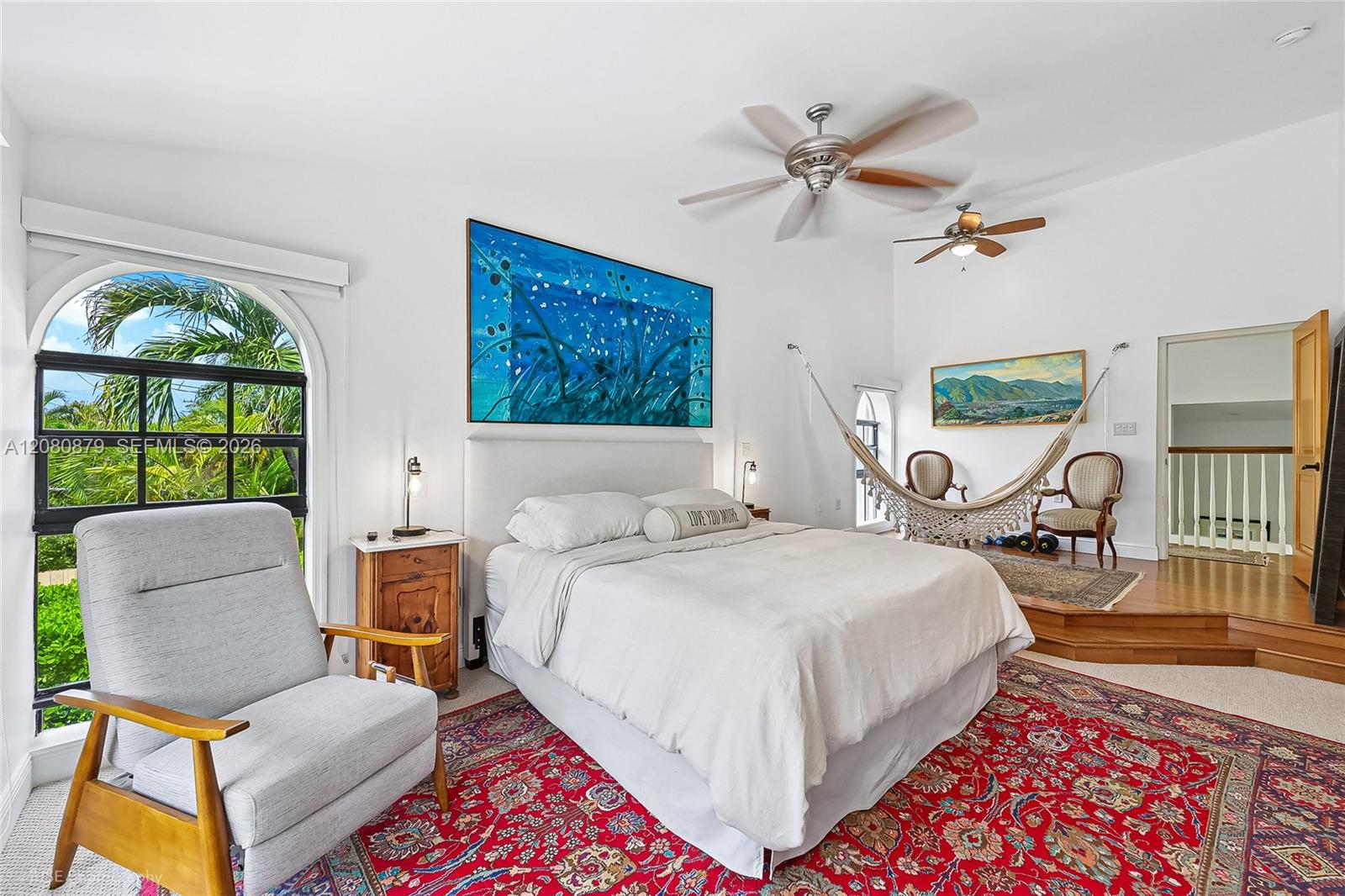 Bedroom with an arched palm-view window, white bed, patterned rug, and hammock.