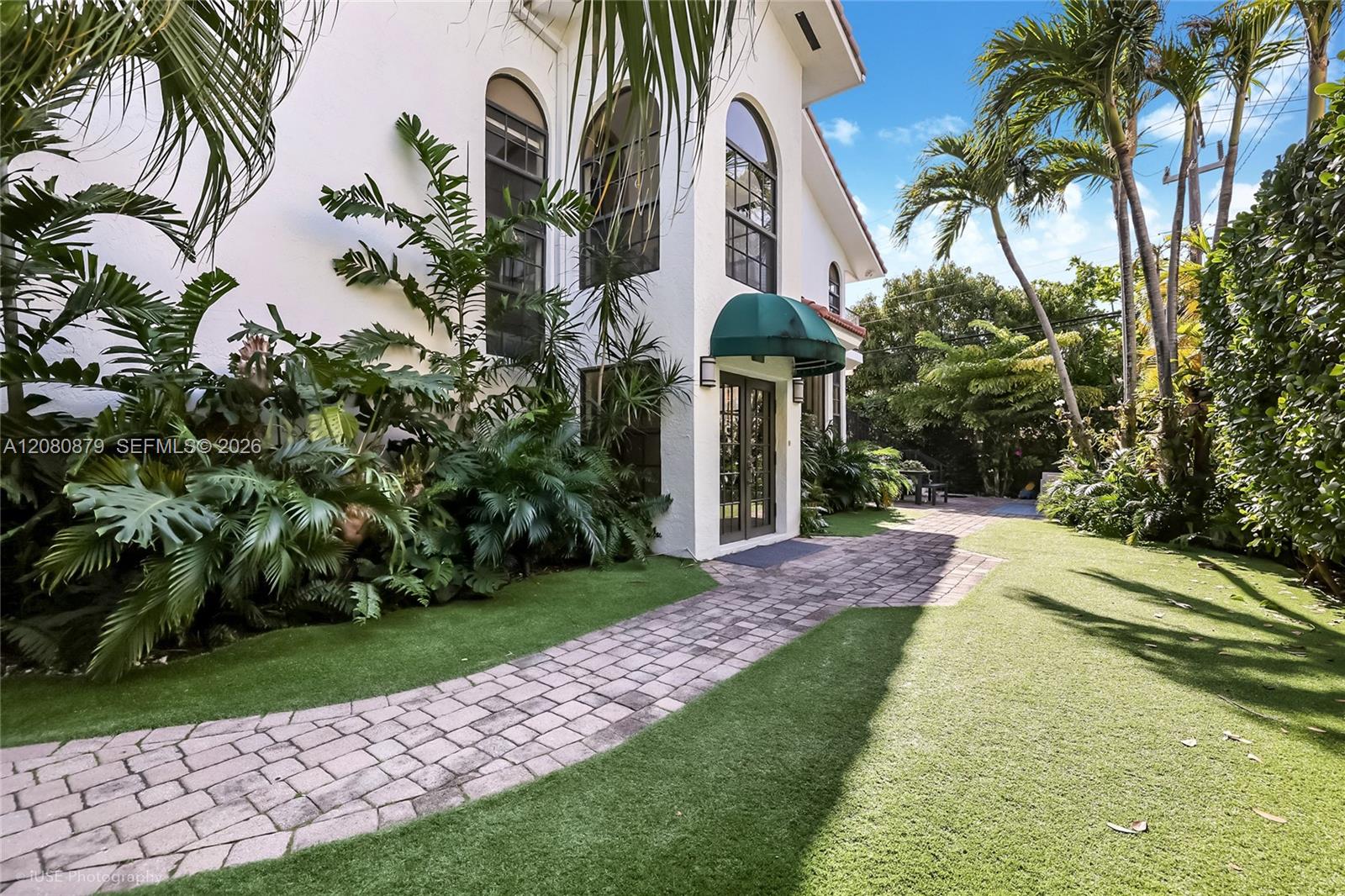 White home with arched windows and a green awning, framed by palms and a curved stone walkway.