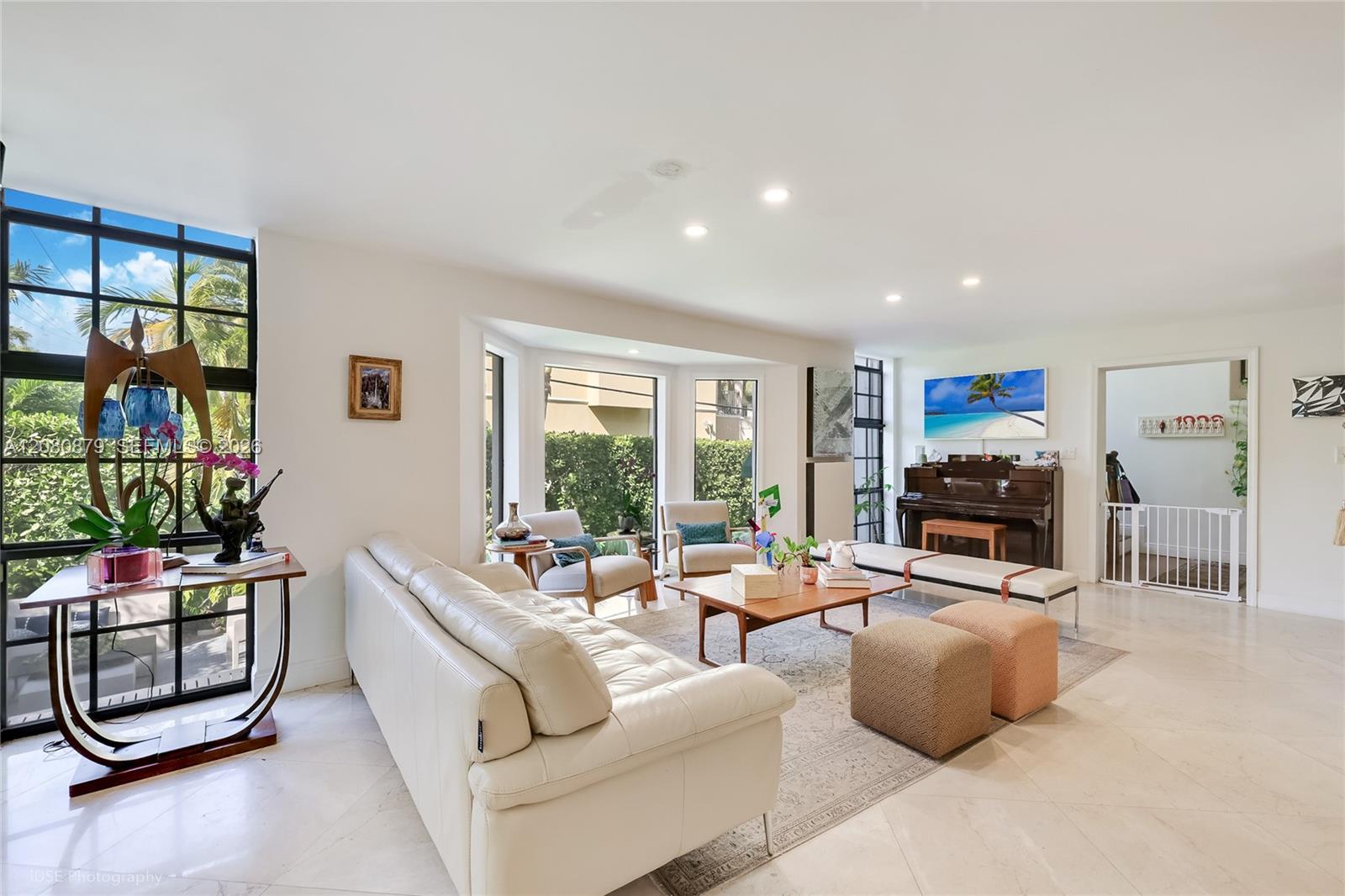 Bright living room with a white leather sectional, large tropical-facing windows, and a piano.