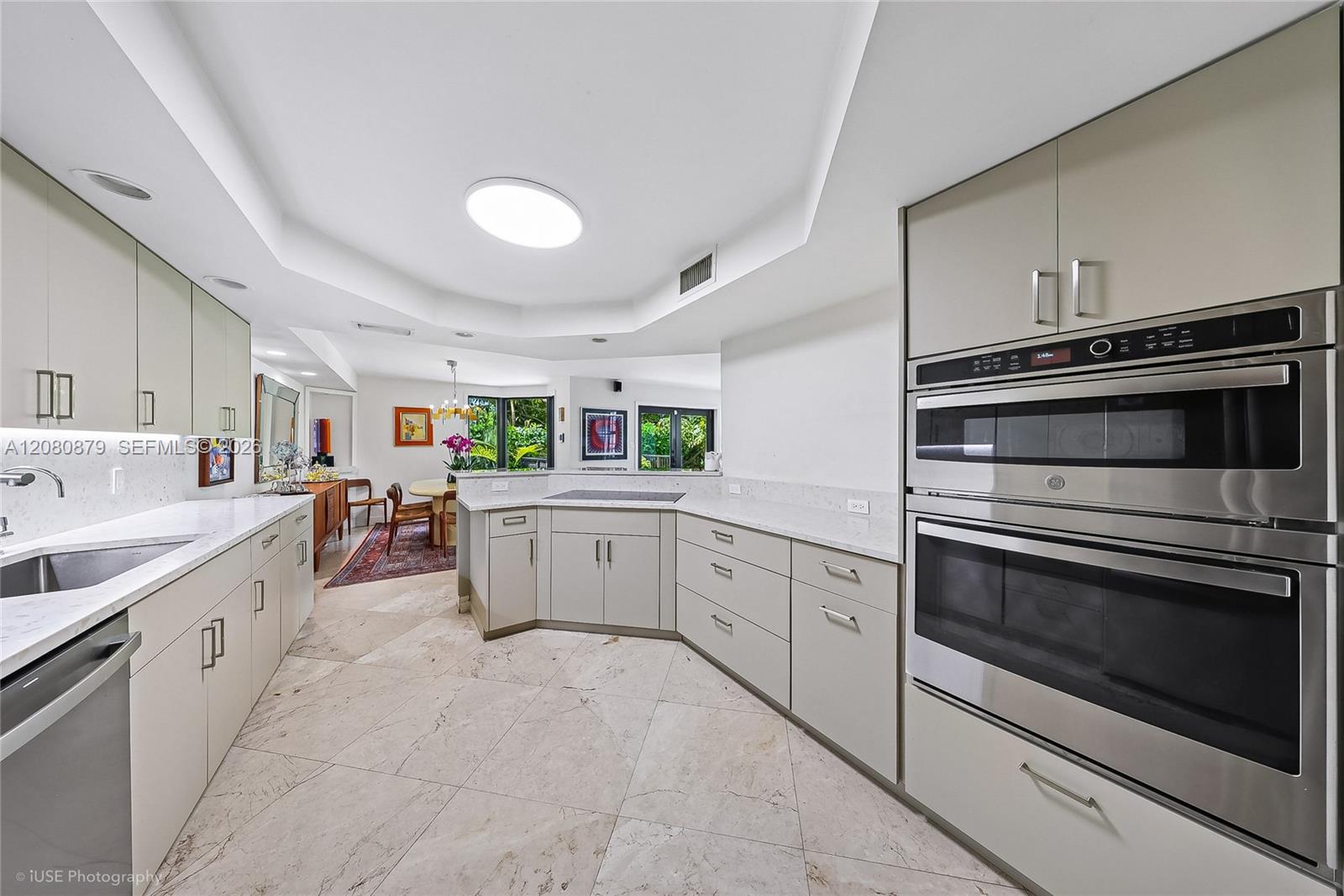Modern kitchen with white stone counters, stainless-steel double ovens, and windows overlooking greenery.