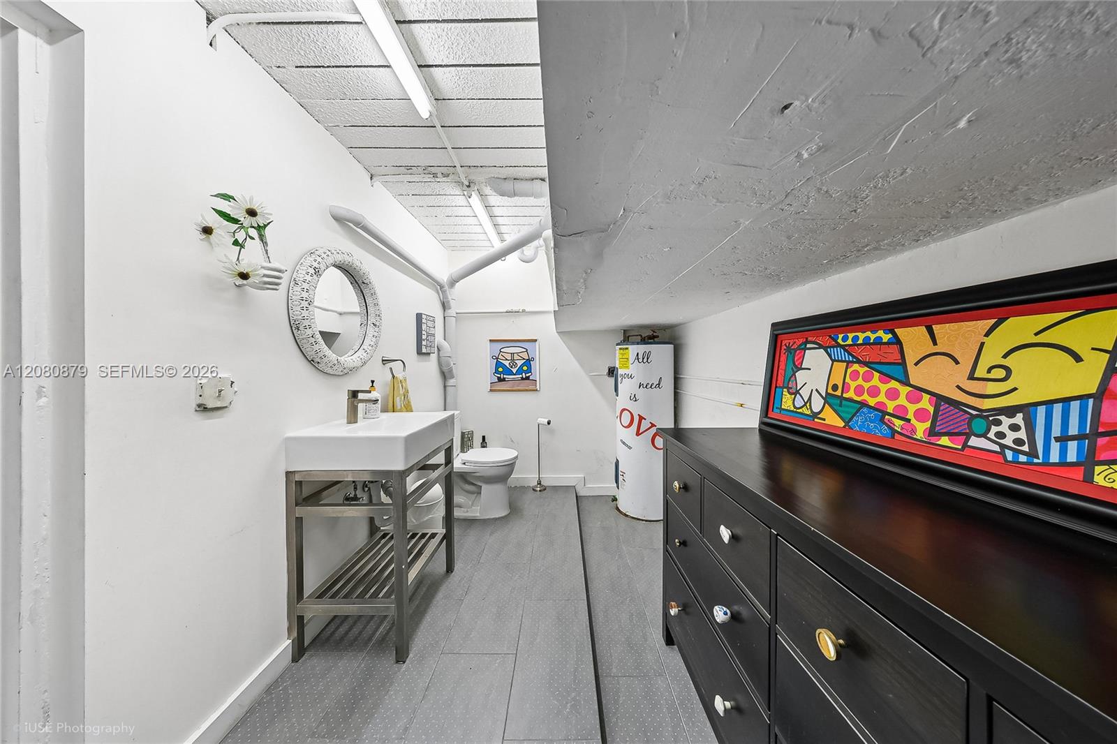 Narrow bathroom with a stainless-steel sink stand, toilet, gray tile floor, and colorful artwork above a dark dresser.