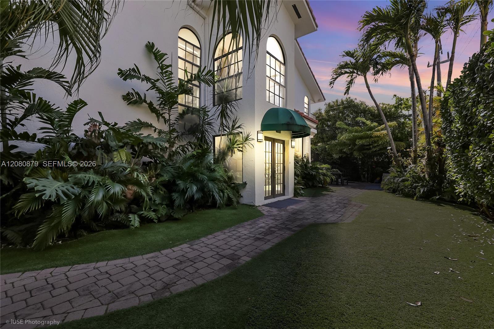 White two-story home with arched windows, a green entry awning, and a curved paver walkway at dusk.