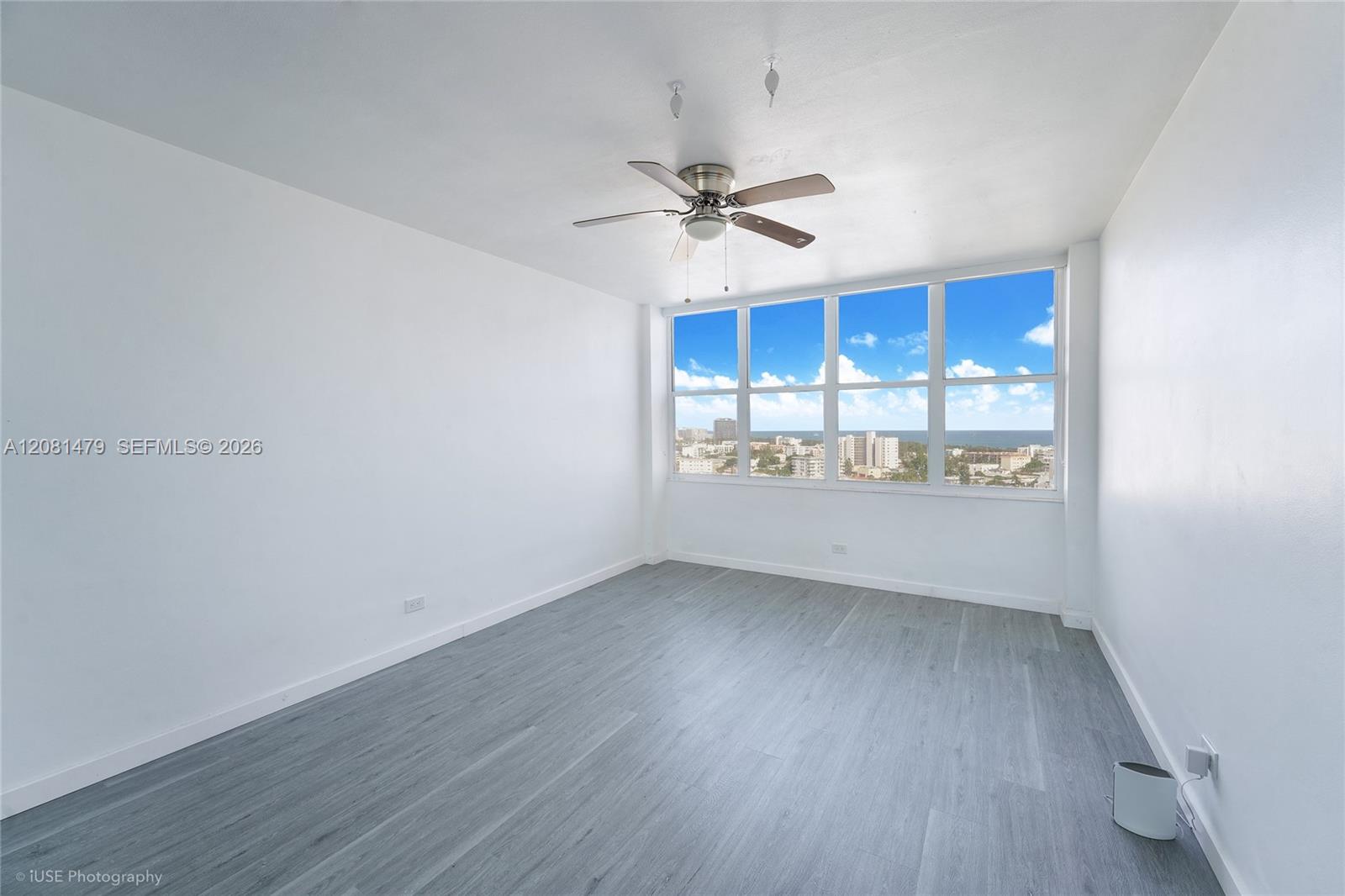 Vacant room with gray plank flooring, ceiling fan, and large windows overlooking buildings and ocean.