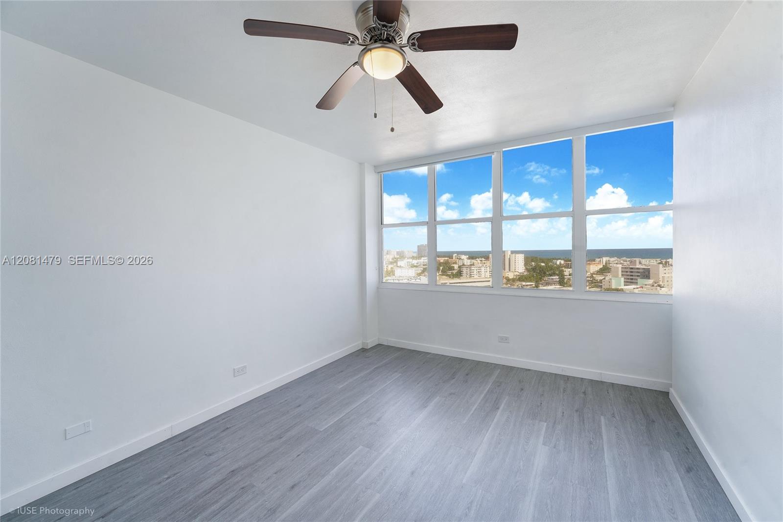 Vacant room with gray flooring, ceiling fan, and large windows overlooking the city and ocean.