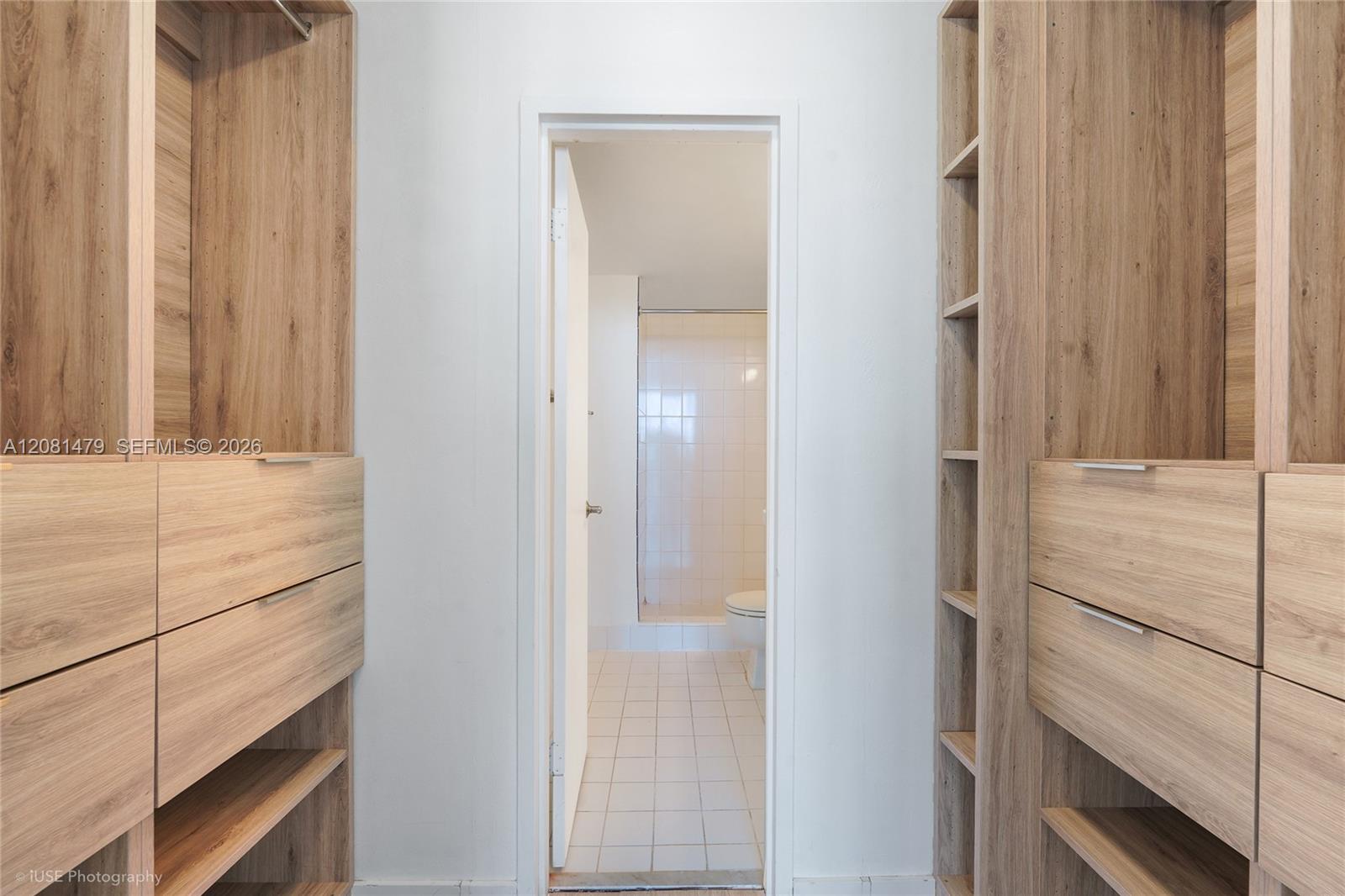 Walk-in closet with light wood built-ins and drawers, opening onto a tiled bathroom.