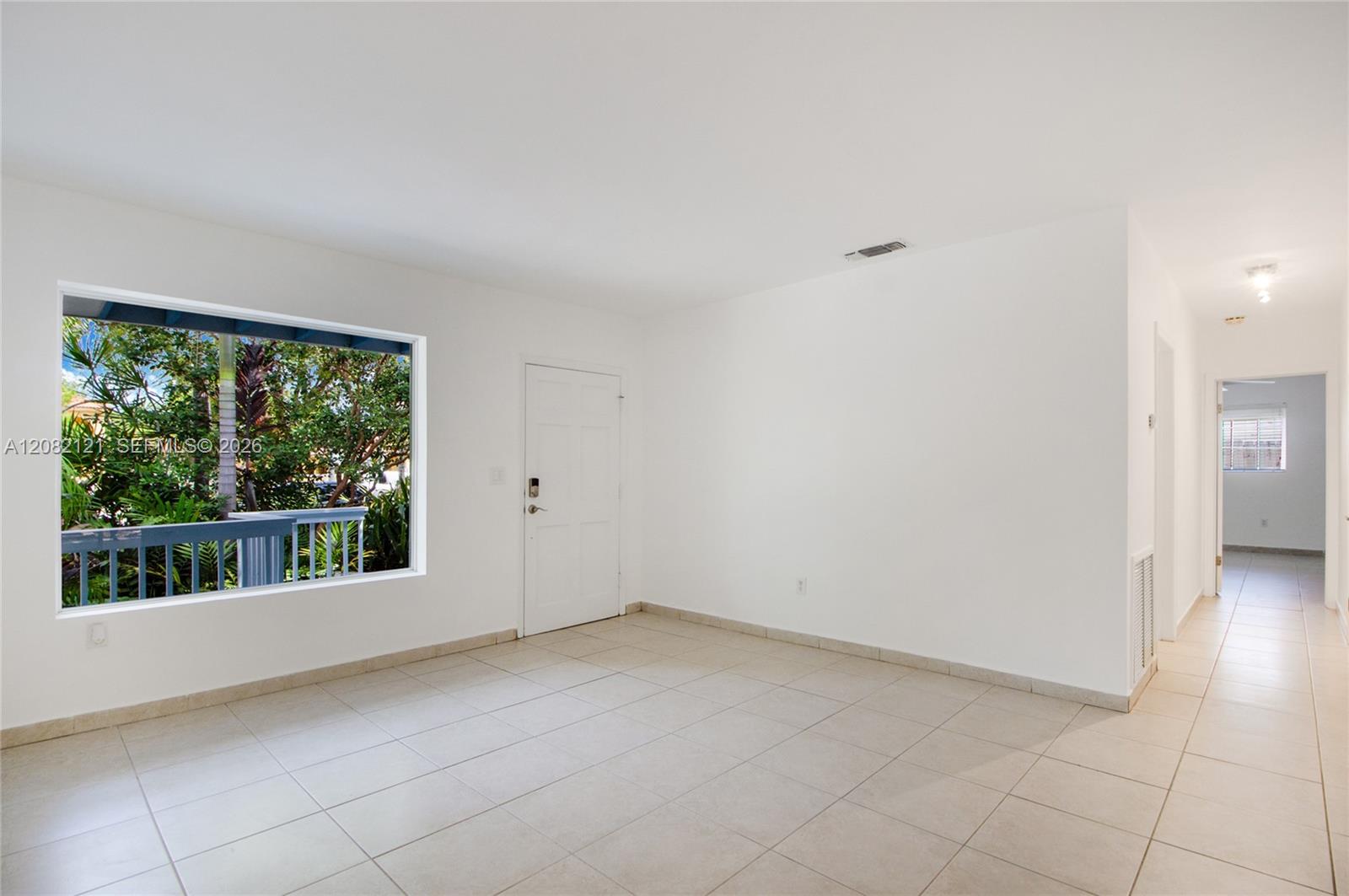 Empty tiled room with a large window overlooking tropical greenery and a hallway.