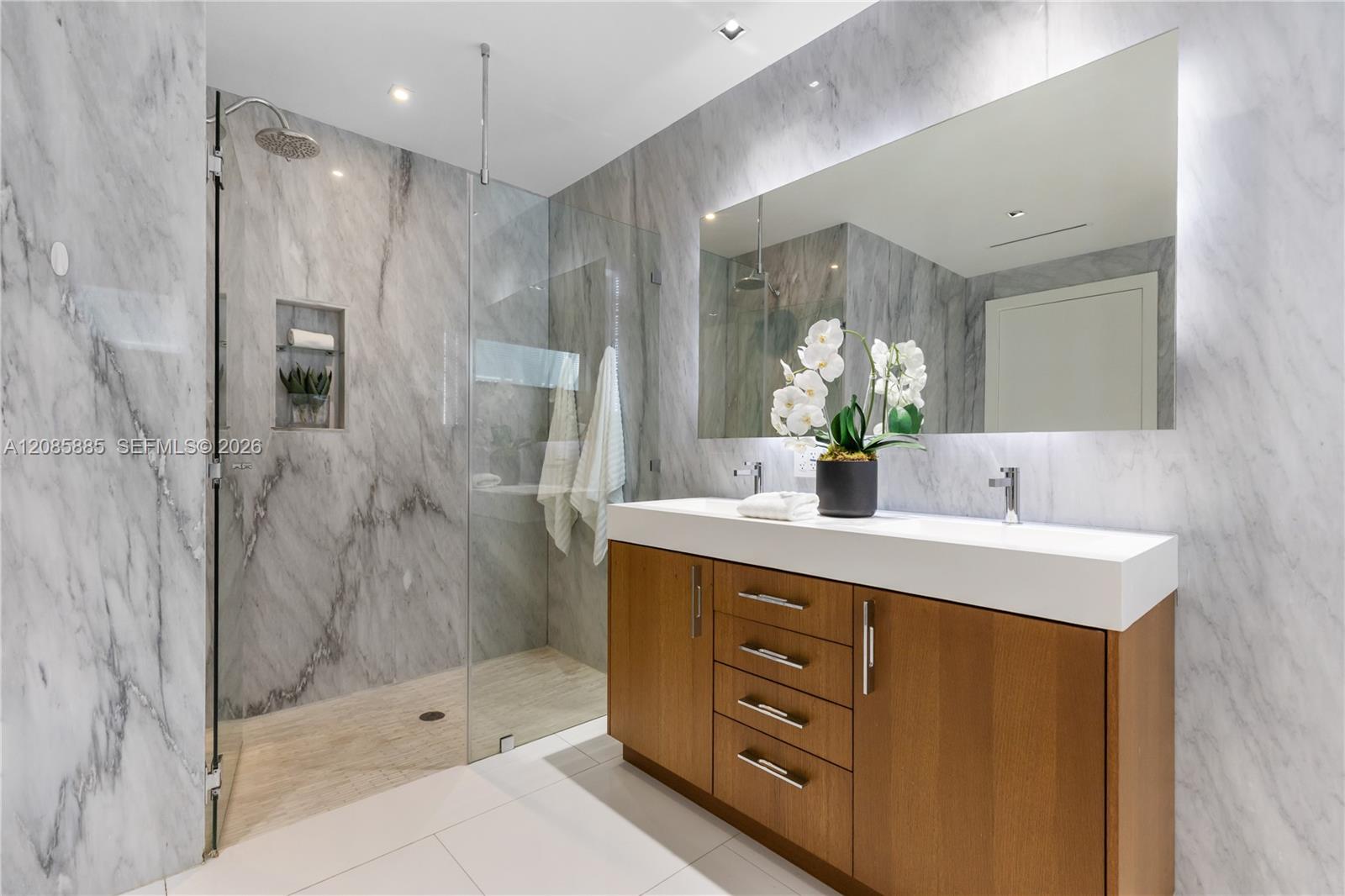 Marble bathroom with a glass walk-in shower and wood vanity topped with white orchids.