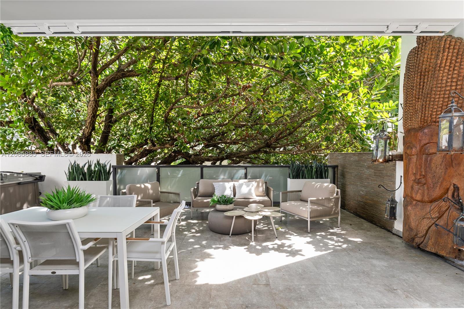 Covered patio with dining and lounge furniture beneath leafy trees and a carved wooden feature.
