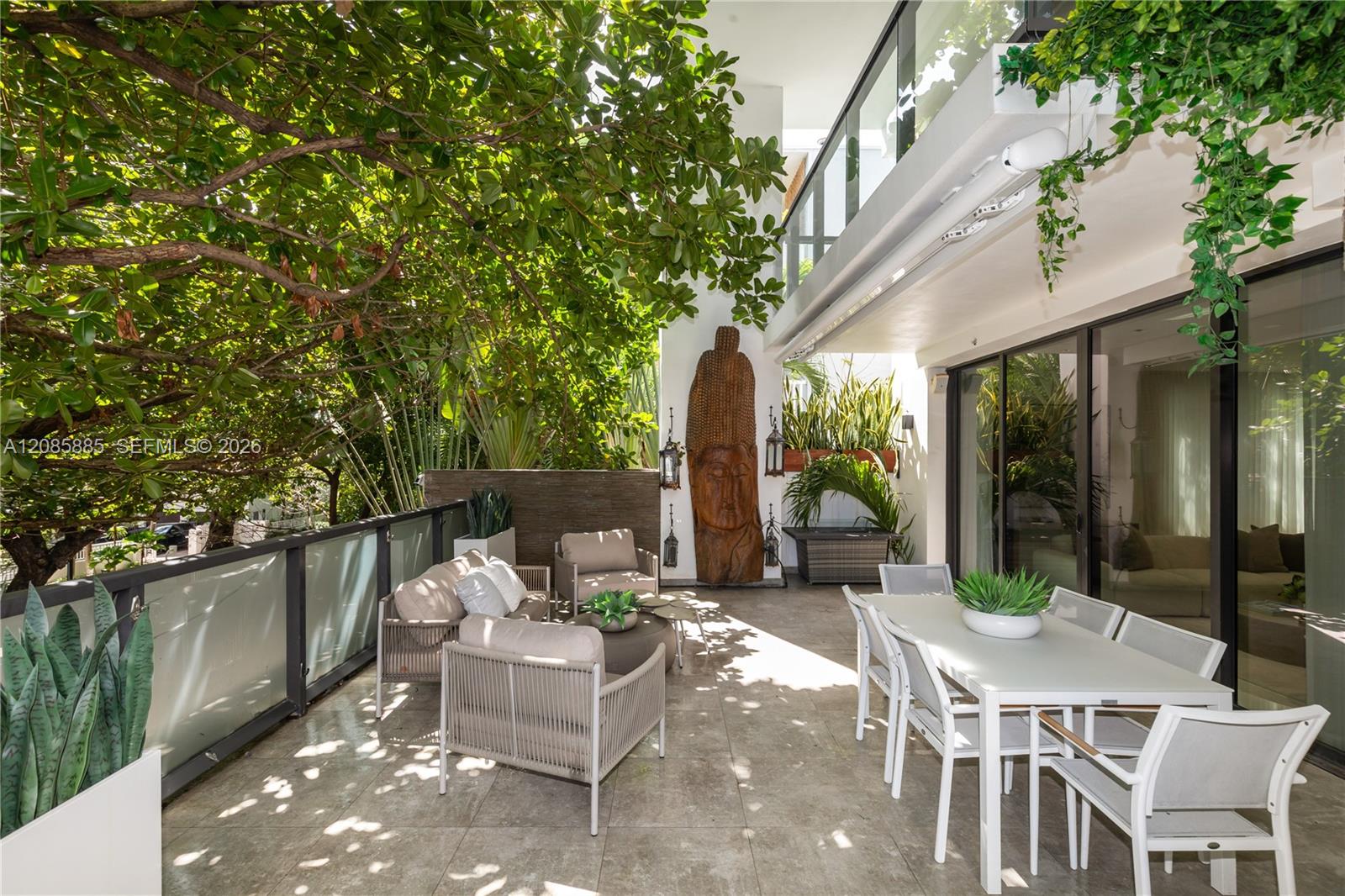 Furnished condo terrace with dining set, lounge chairs, leafy canopy, and carved wooden figure.