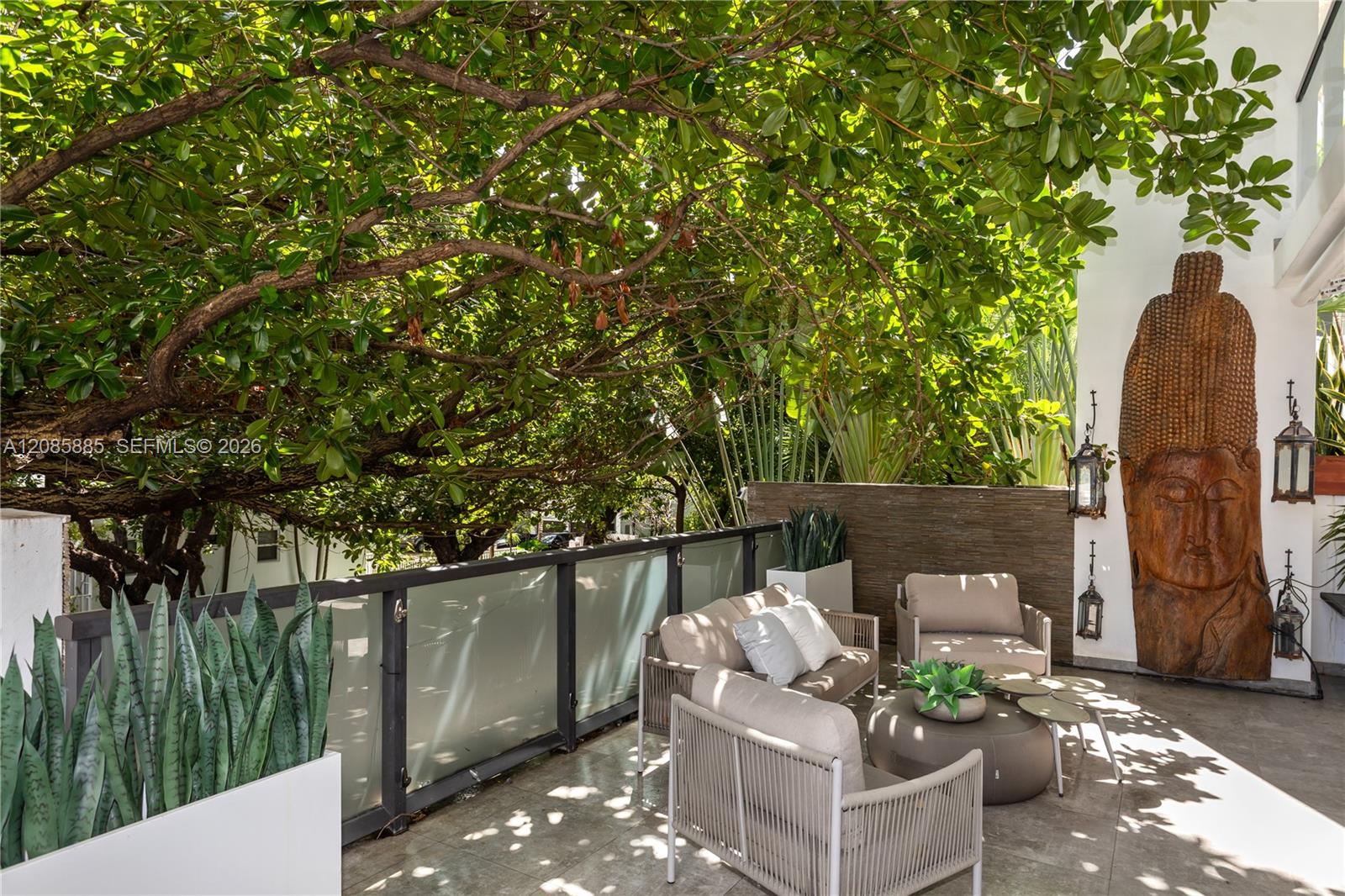 Furnished patio beneath a broad tree canopy, with tropical plants and a carved wooden statue.