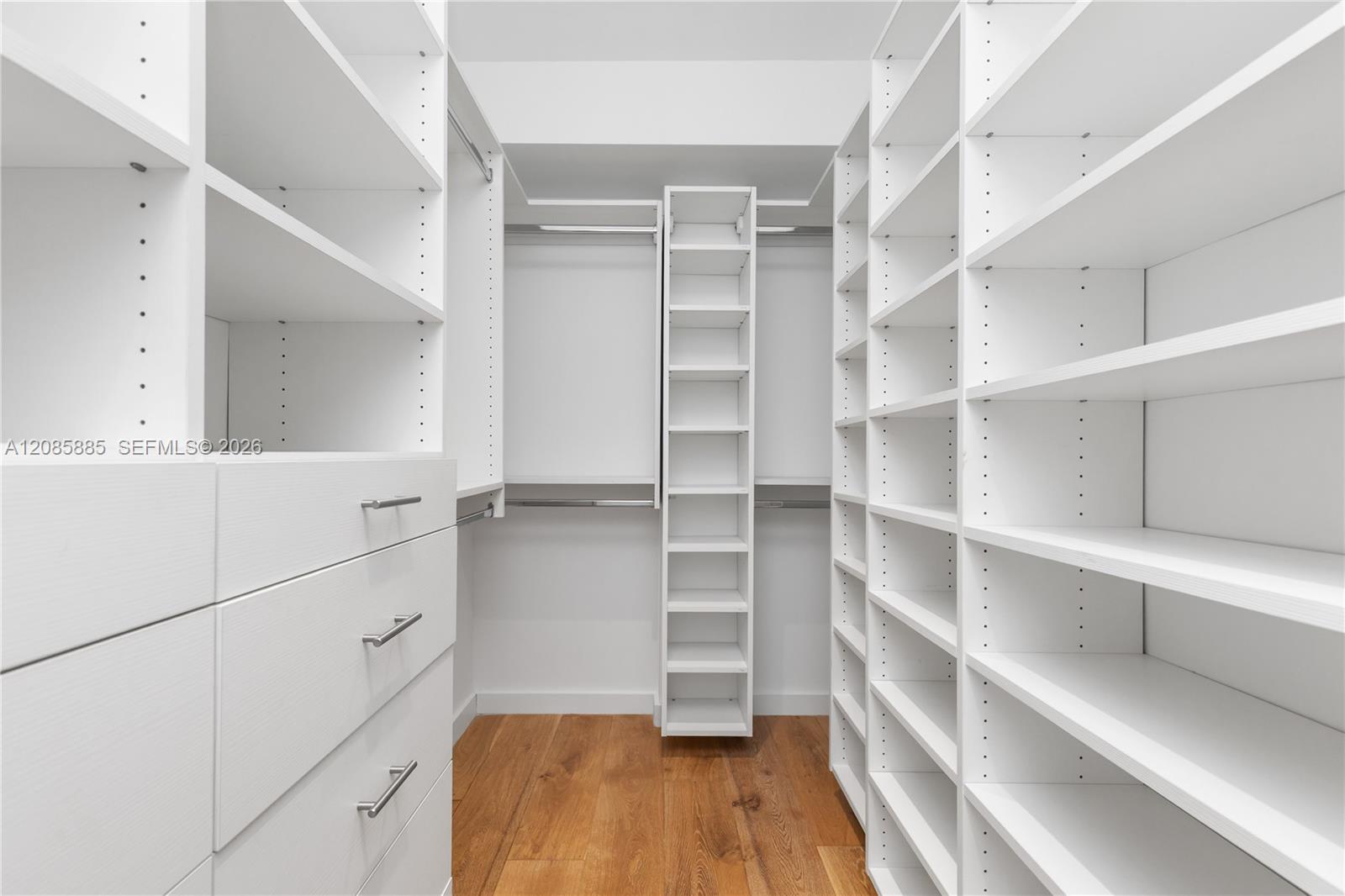 Walk-in closet with white built-in shelves, left-side drawers, and warm wood flooring.