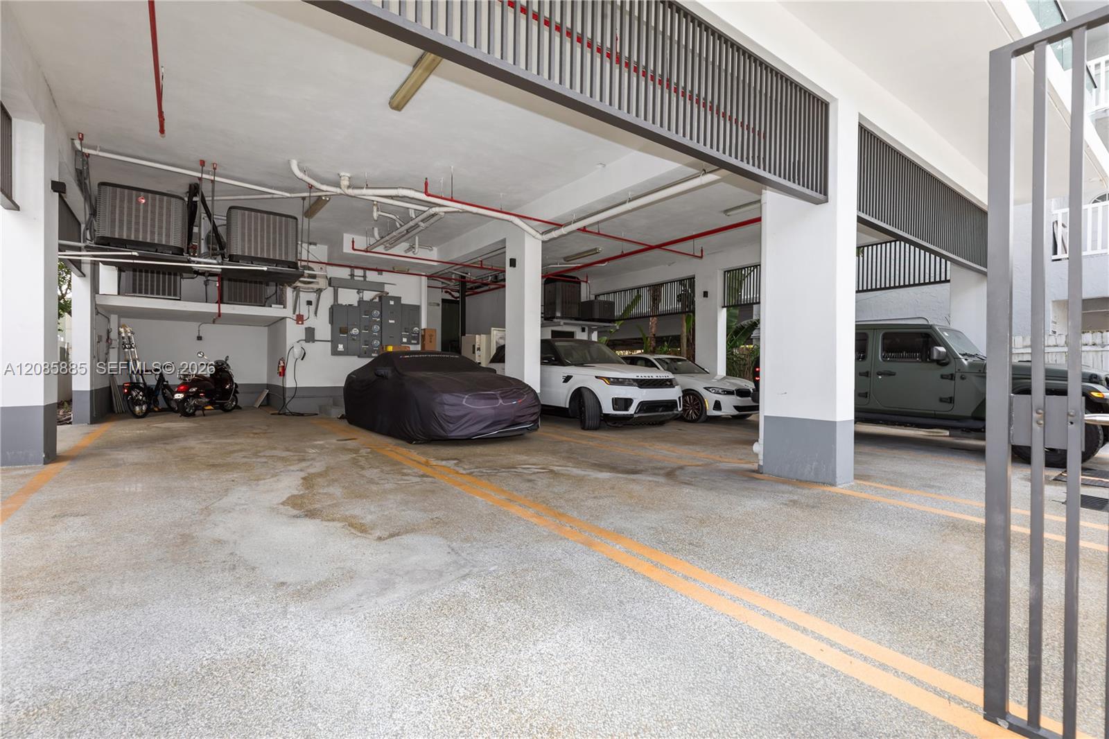 Covered parking garage with marked spaces and several vehicles beneath exposed pipes and ventilation equipment.