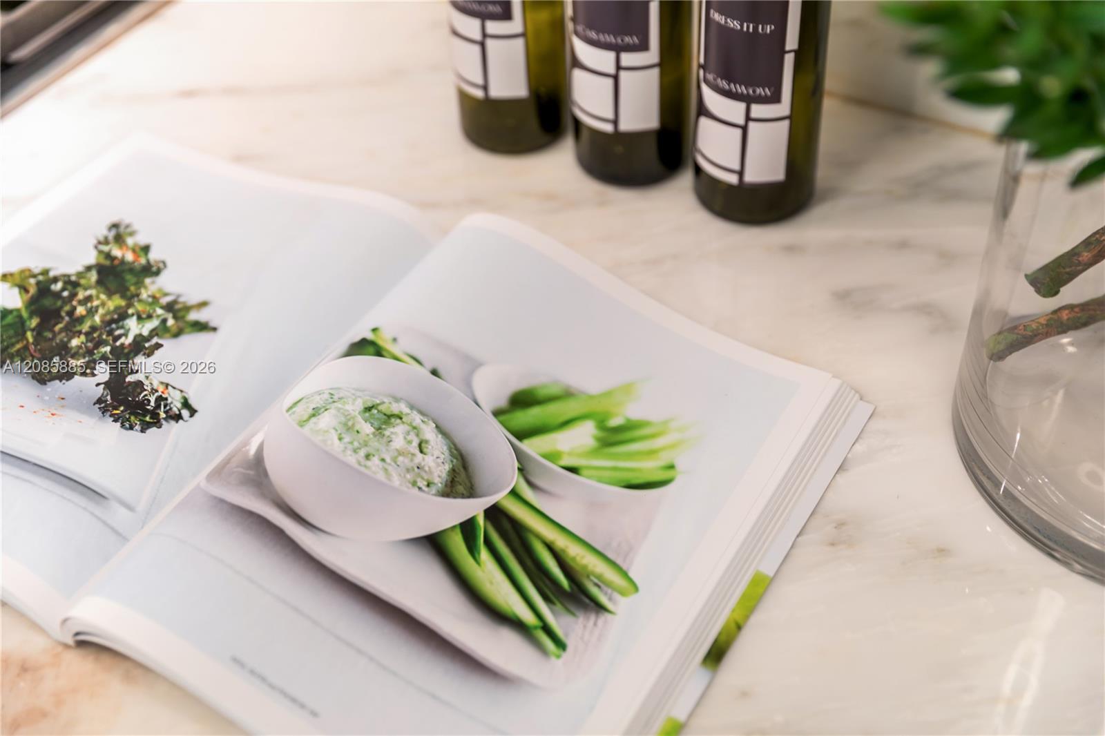Open cookbook with green vegetable dishes beside dark bottles on a light marble countertop.