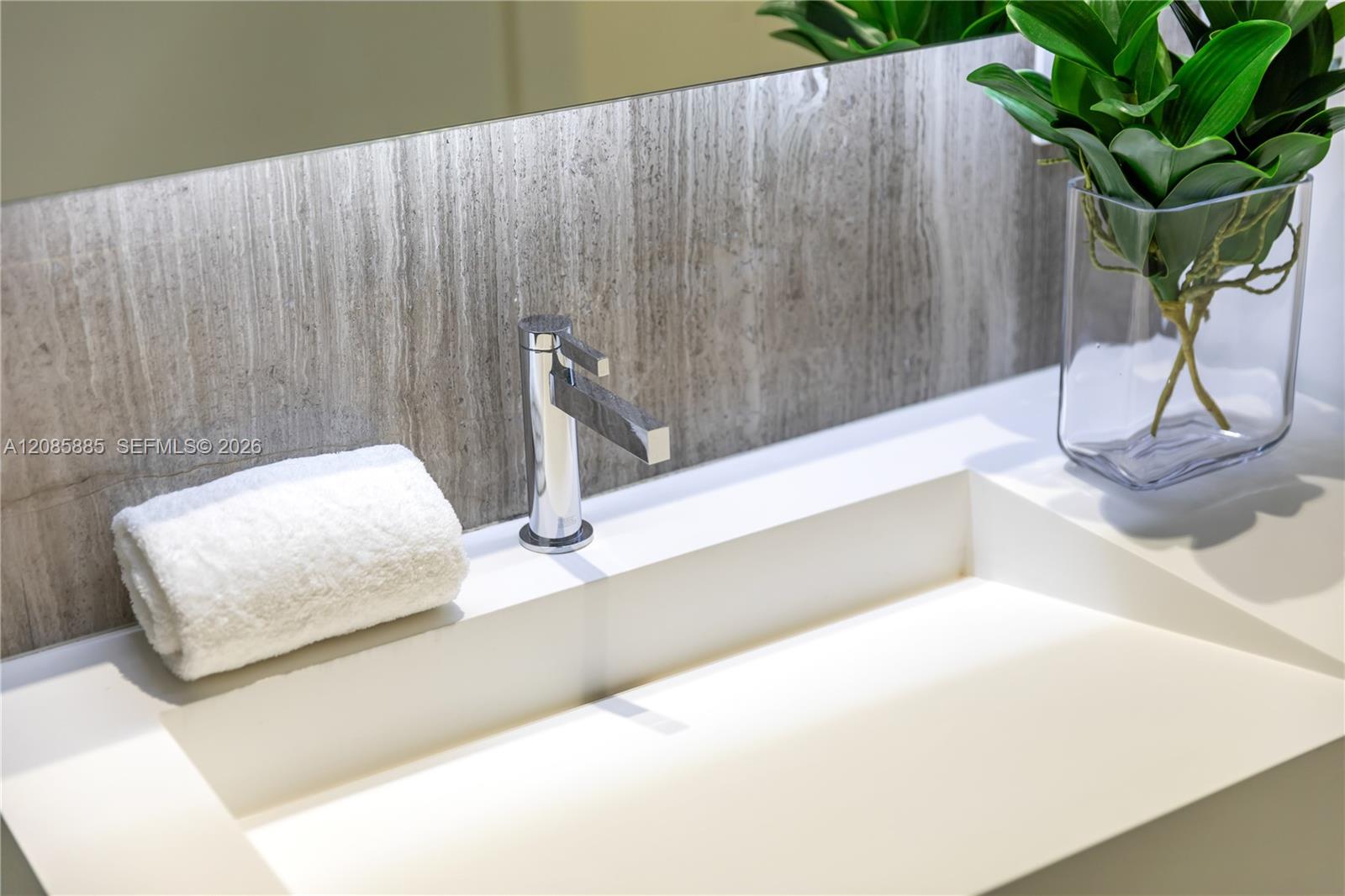 White bathroom sink with chrome faucet, gray stone backsplash, folded towel, and leafy plant.